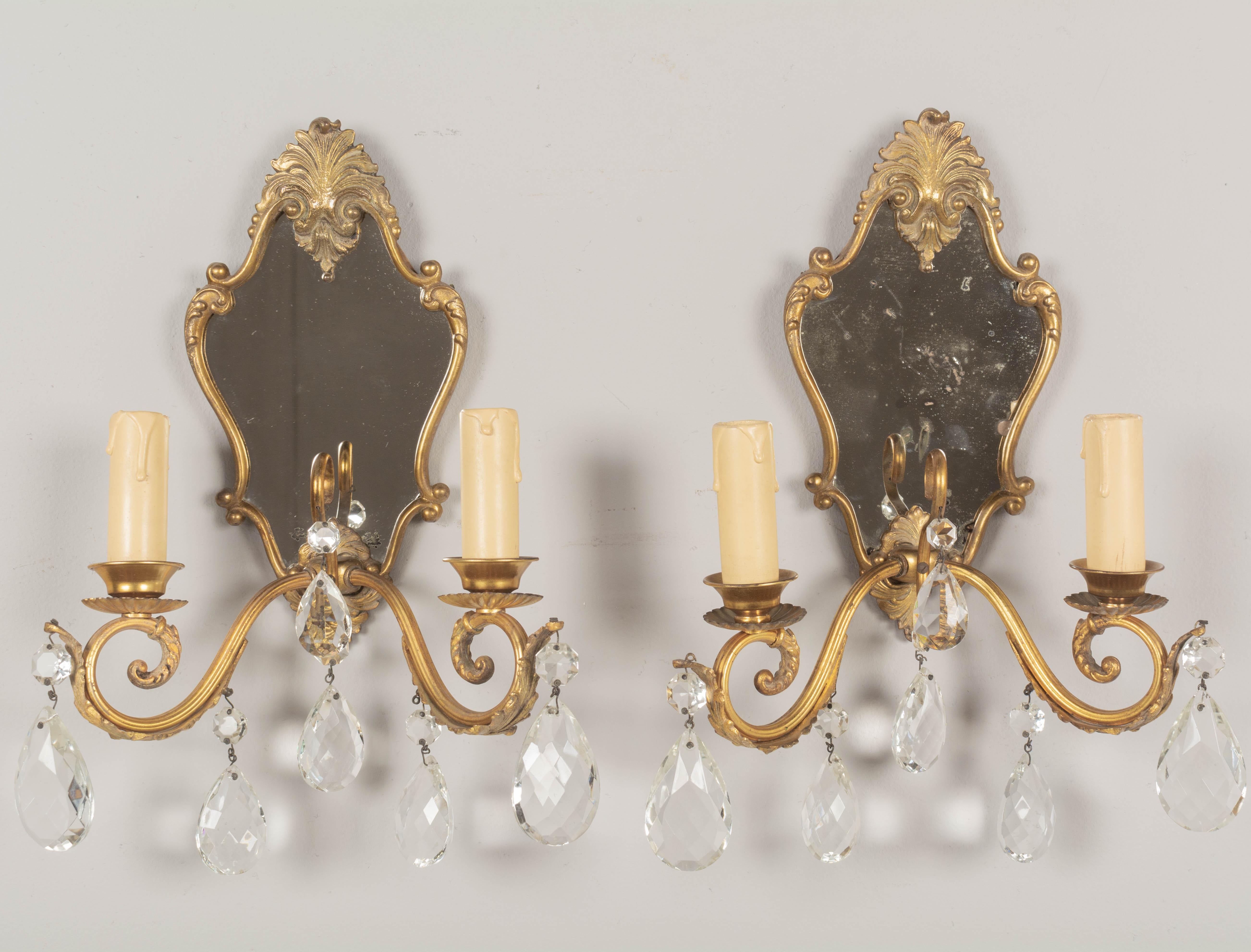 A pair of Louis XV style French cast brass and crystal sconces with mirrored backplates. Good quality heavy casting with two scrolled candle arms and cut crystal teardrop pendalogues with faceted beads. Original candle covers. Rewired with new