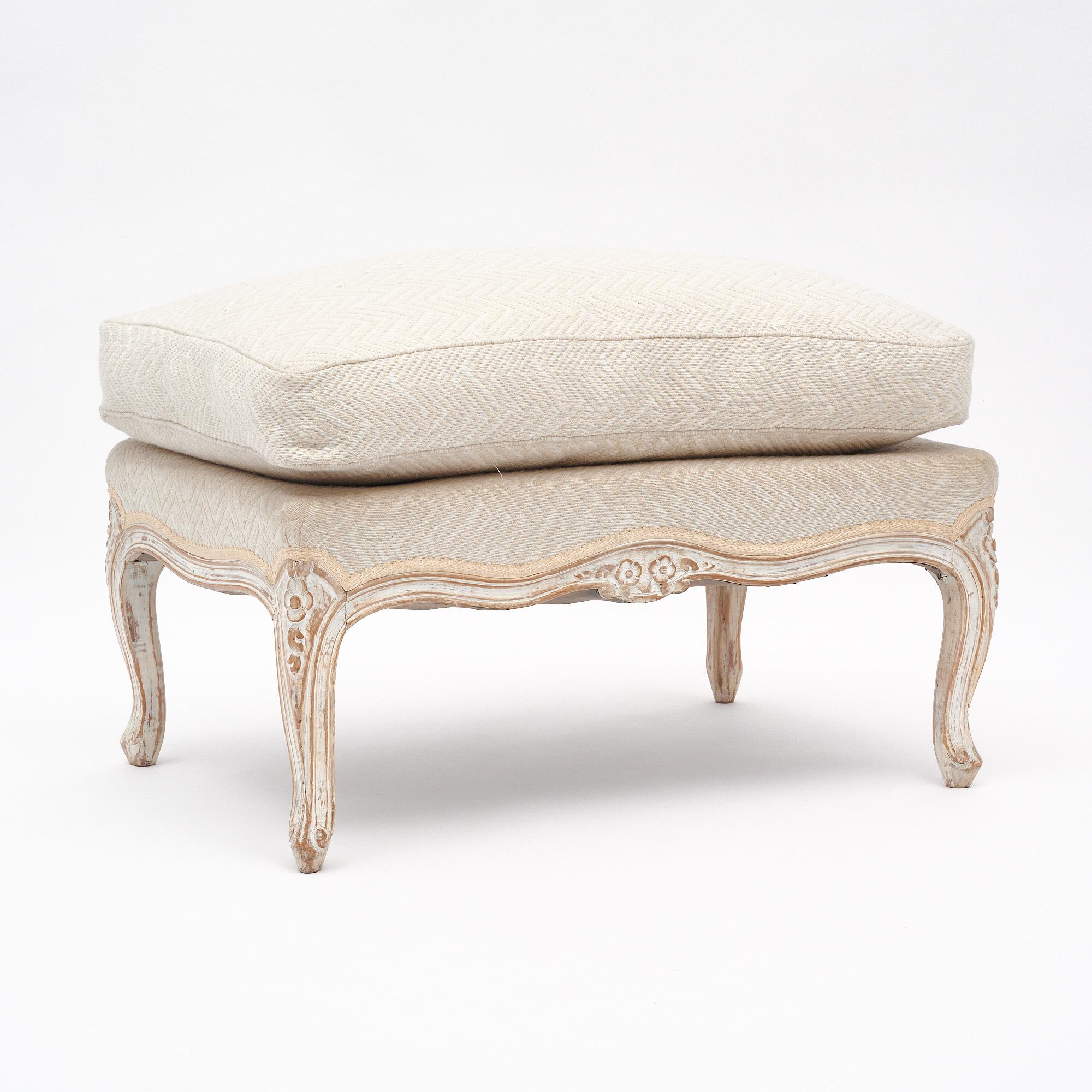Ottoman from France in the Louis XV style. This piece features a hand-carved wood frame with original white paint and patina. The carving includes floral detailing. The upholstery is newly done in a white fabric.
