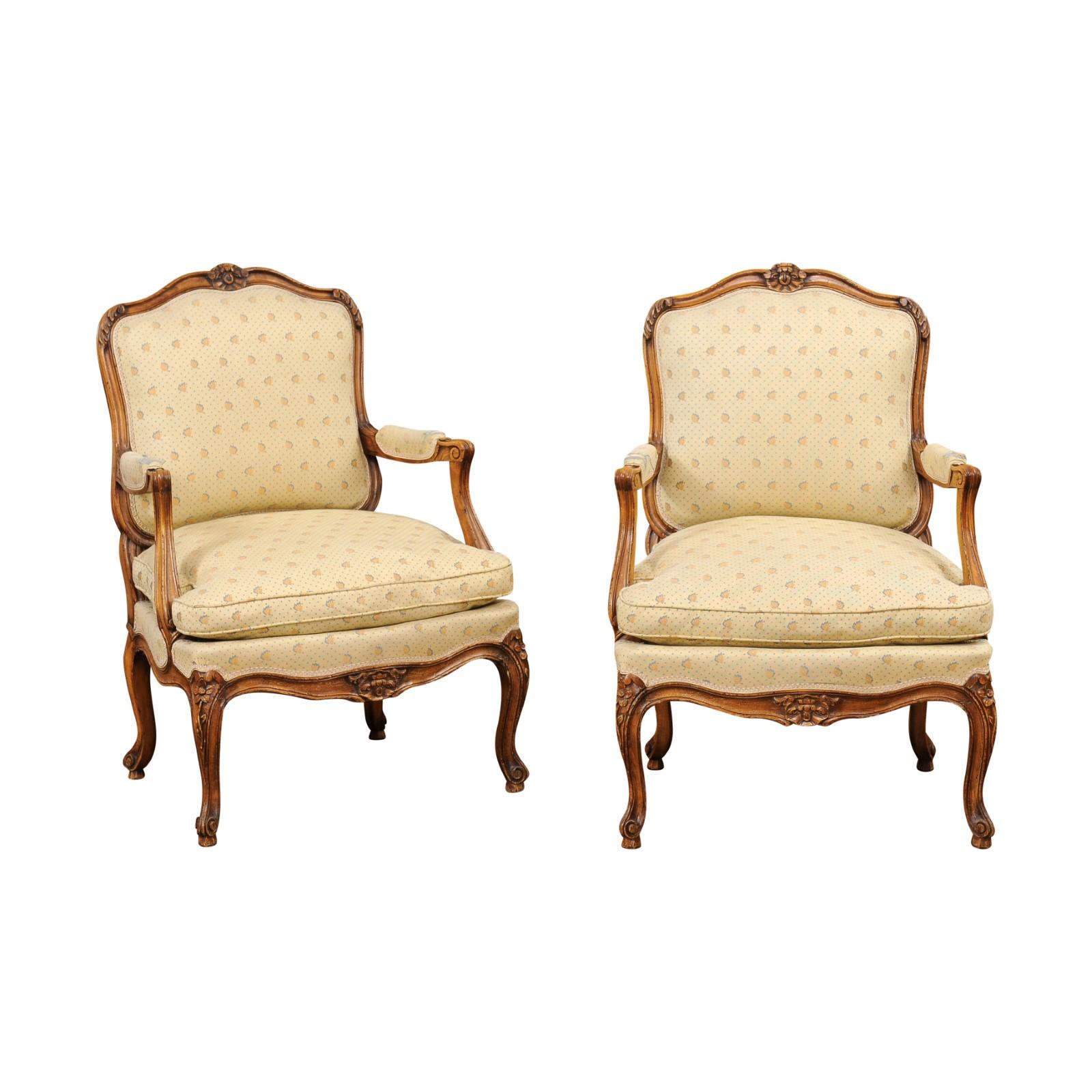 A pair of French Louis XV style walnut armchairs from the 20th century with carved floral décor, scrolling knuckles, cabriole legs and upholstery. Evoking the grandeur of French royalty, this exquisite pair of Louis XV style walnut armchairs, or