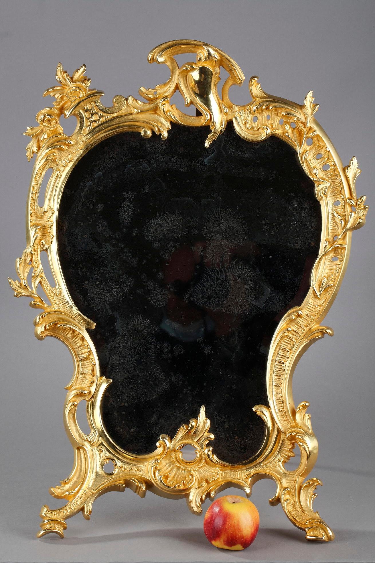 This ornamental Louis XV style mirror glass captures the grace of the Rococo age. Crafted of gilt bronze, the frame features sweeping, heavy scrolls and acanthus with floral details. A singing bird adorns the top of the golden frame. The back part