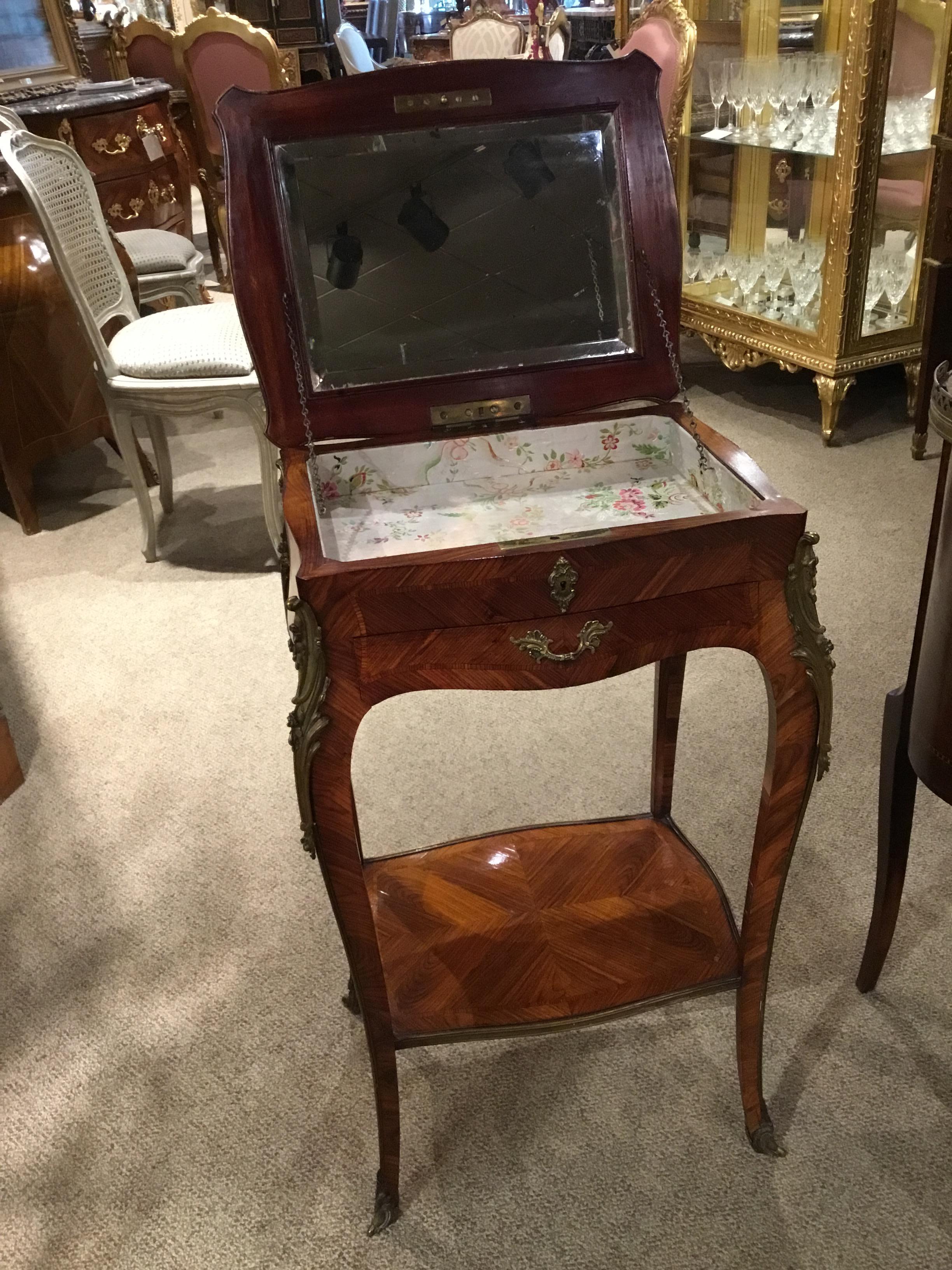 Beautifully shaped petite table with a top that lifts open to a recessed area with a beveled mirror.
Has a single drawer and a shelf on the lower tier. Has antique bronze mounts on each leg.
