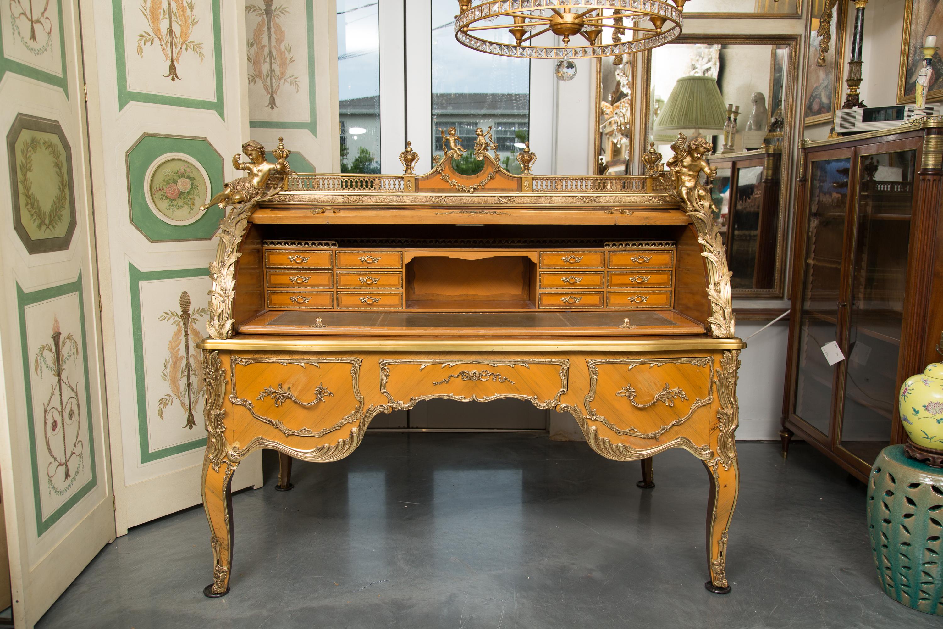 This is a stately and lavishly decorated Louis XV style cylinder desk. The top with a pierced brass gallery centered by an inverted arched pediment with cherubs. The top is flanked by four prominent brass figural putti and candle holders above an