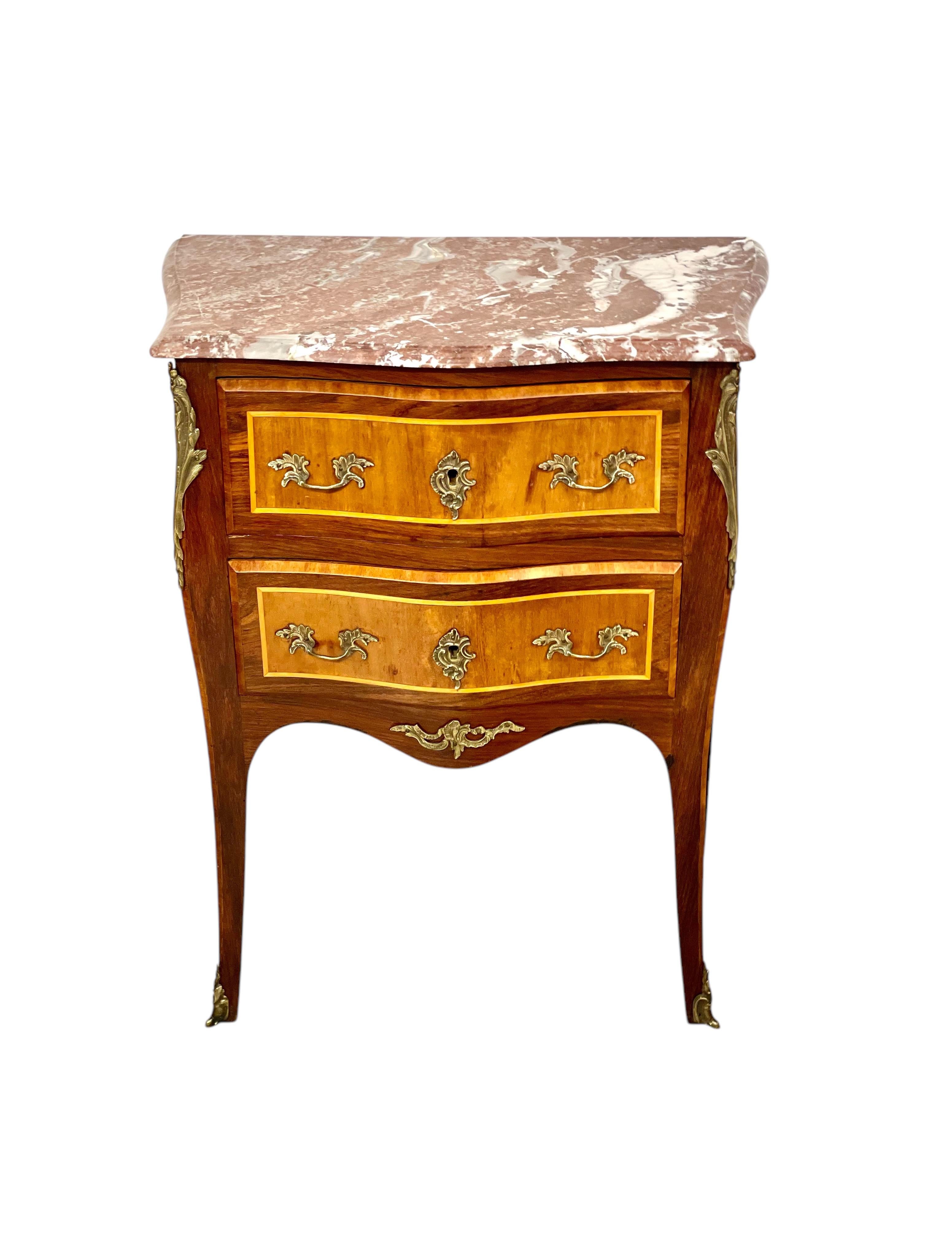 Questa piccola commode è un affascinante esempio di artigianato del XIX secolo ispirato al classico stile Luigi XV. Realizzato in legno impiallacciato e splendidamente lavorato, il comodino presenta curve aggraziate su tutti i lati. Si apre per