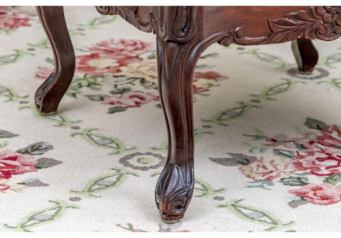 A Louis XV style square upholstered bench with a dark, richly carved walnut wooden frame with ornate details on the legs and scalloped apron. The bench upholstered in a textured woven blue tonal fabric with double welting and resting on cabriole