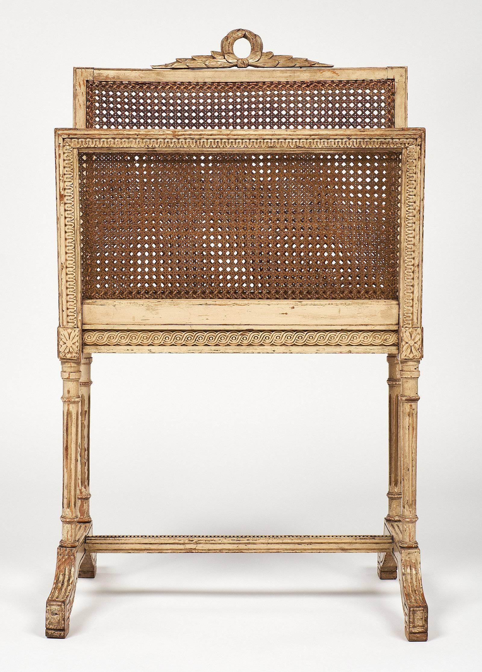An elegant, neoclassical style magazine rack with its original ivory color paint and patina. This piece is made of beech wood. The caning is original as well and in beautiful condition, with finely carved floral details and laurel crown.