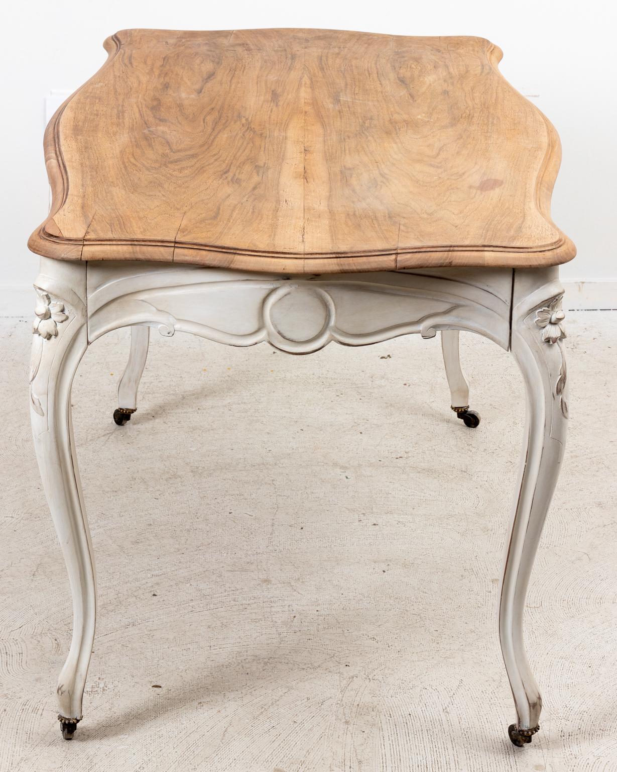 Circa 1940s Louis XV style white painted table with carved flowers on the curved apron and cabriole legs. The base is painted chalky white with a stripped Fruitwood top surface. One drawer opens from one side. The curved legs also end on castors.