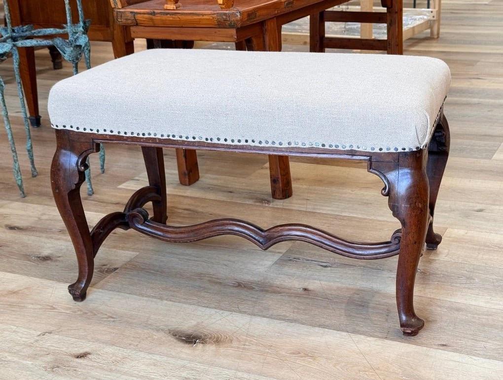 A finely carved Louis XV walnut bench or stool, France, 18th century, featuring cabriole legs, a shaped stretcher, and scrollwork supports. The seat is newly upholstered in natural linen with nailhead trim. Elegant and versatile, it reflects the