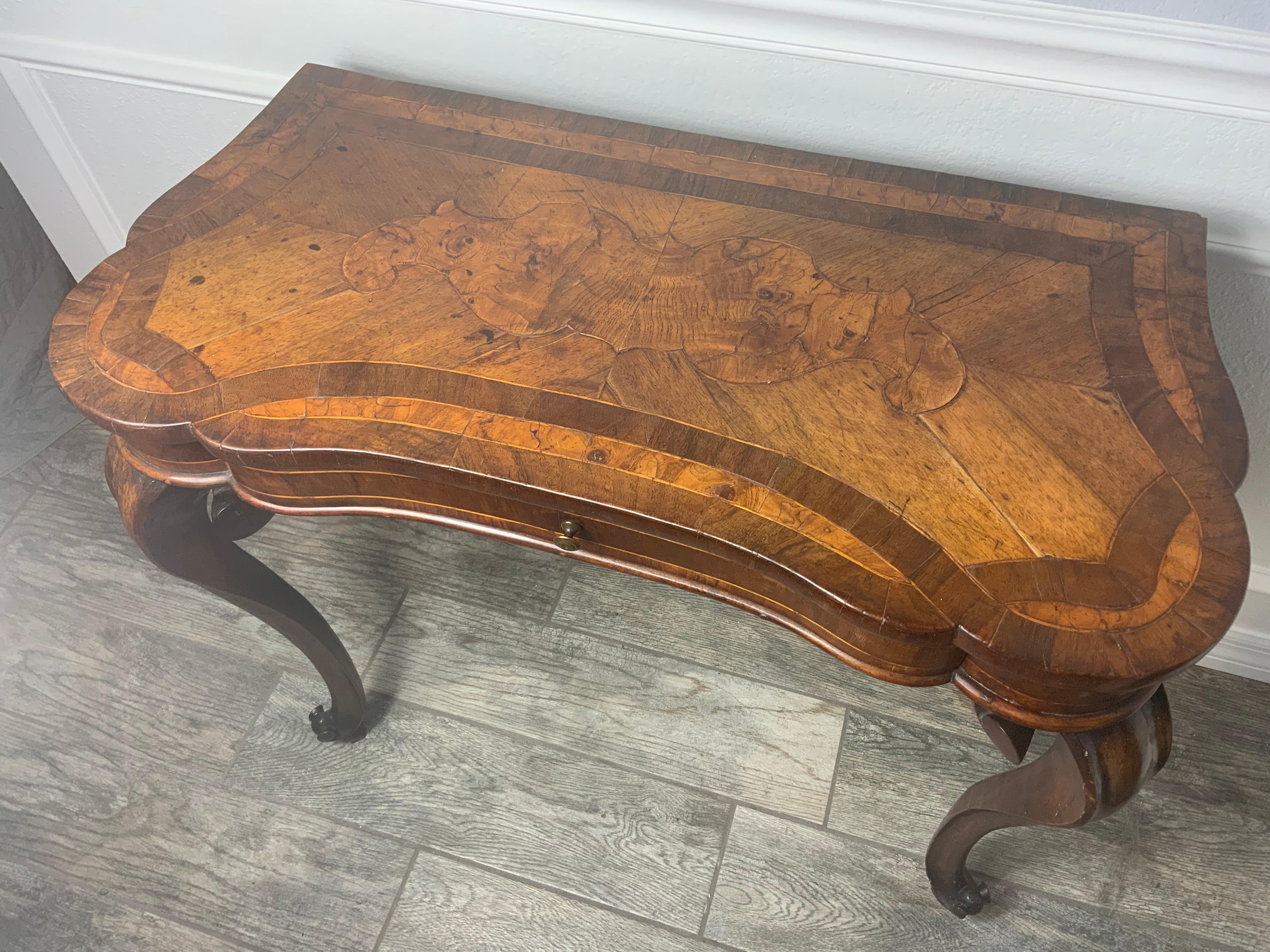 A Queen Anne era Walnut writing table, with bordered edge and book matched burl walnut veneer with mixed wood inlays to the top with rounded corners to the front, shaped inlay apron, raised on large scrolled carved cabriole legs on a raised carved