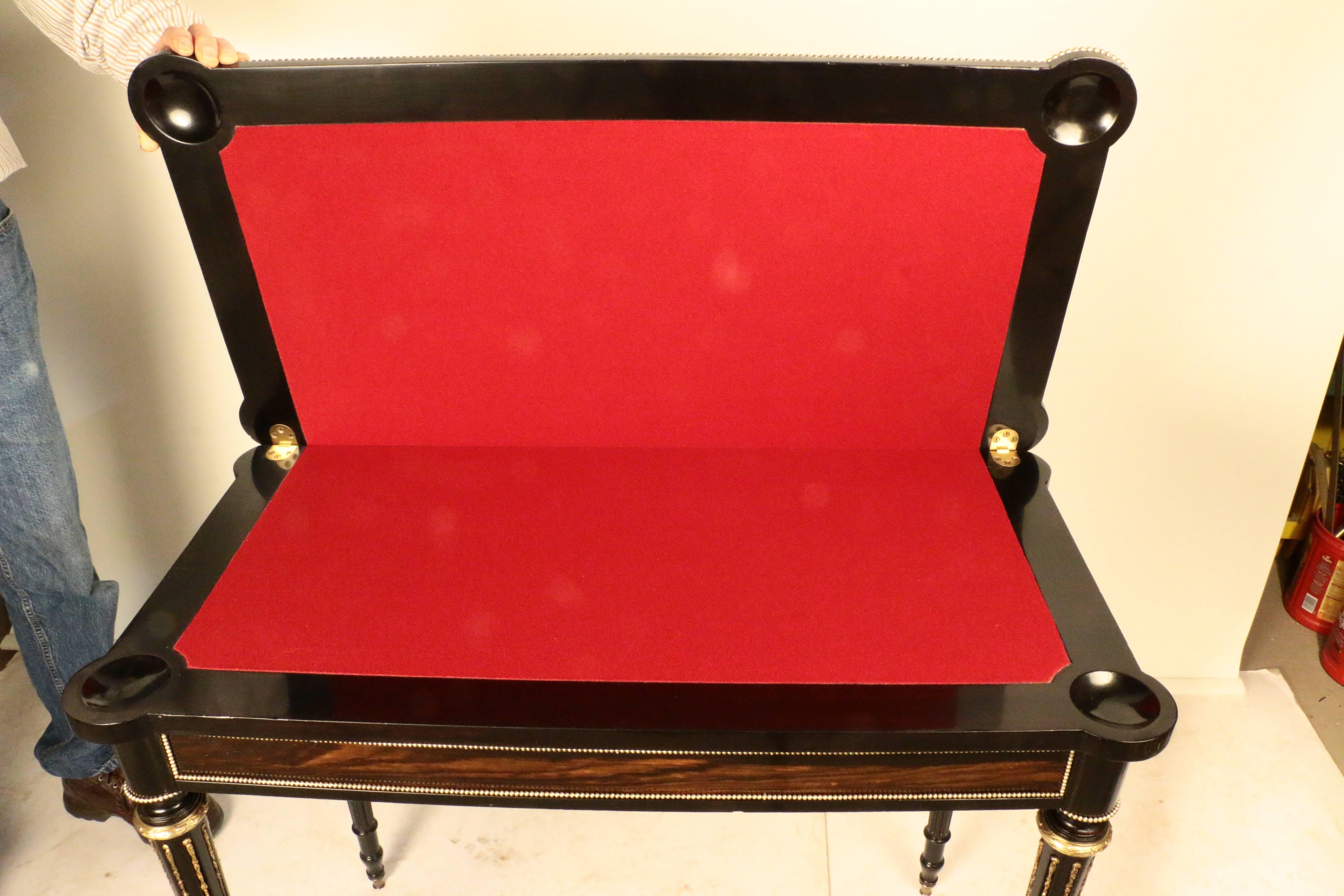 The boldly figured coromandel surface with eared corners, opening to reveal a red baize surface, framed in ebonized mahogany, with a counter -well at each corner. The surface and paneled sides are edged with brass beading, There is a storage area