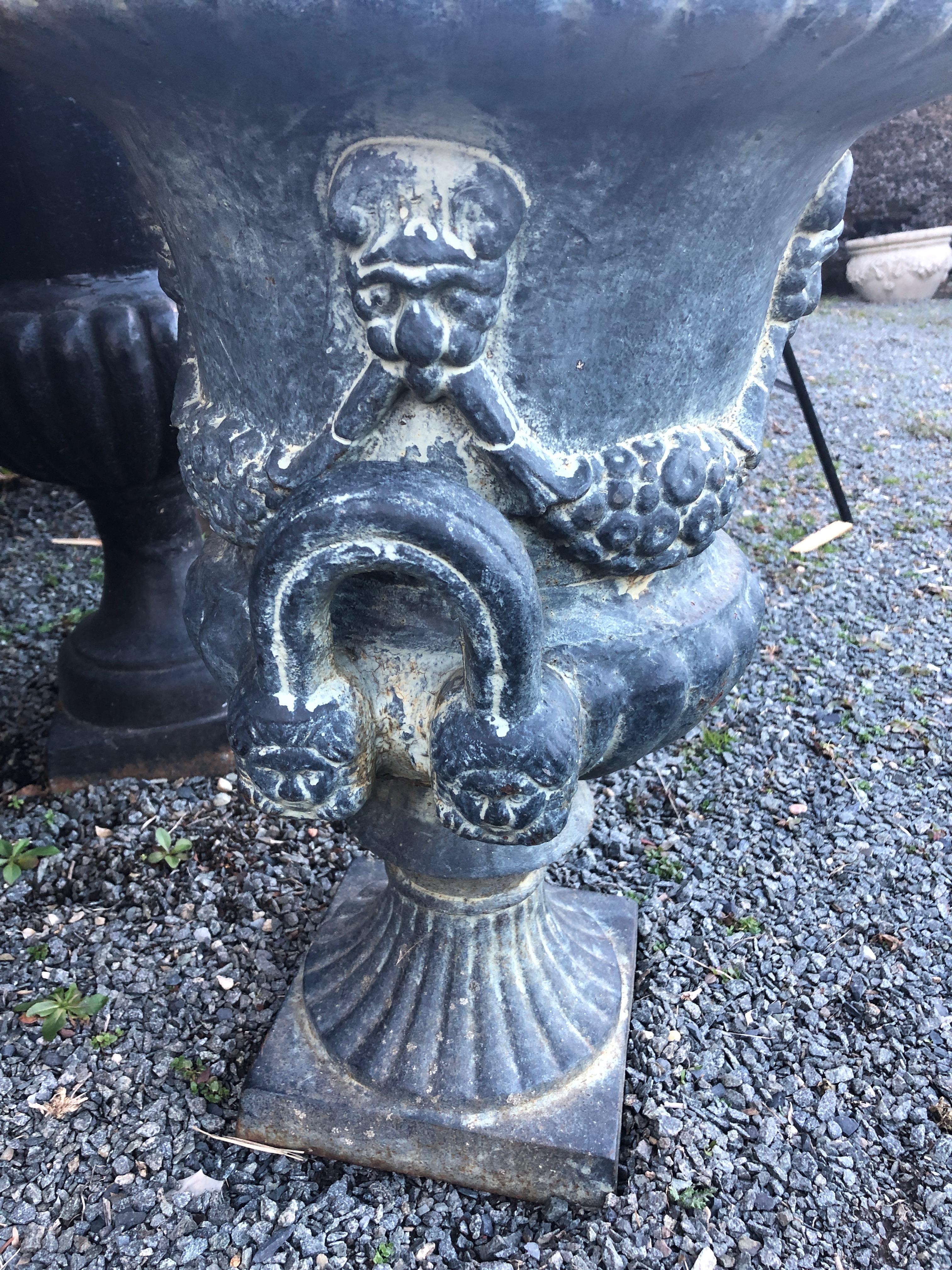 This cast iron jardinière of Classic campana or bell-shaped urn form features a gadrooned edge, raised neoclassical swags and Roman male masks on the body, and handles on either side. Set on a fluted column base with plinth foot, its natural patina