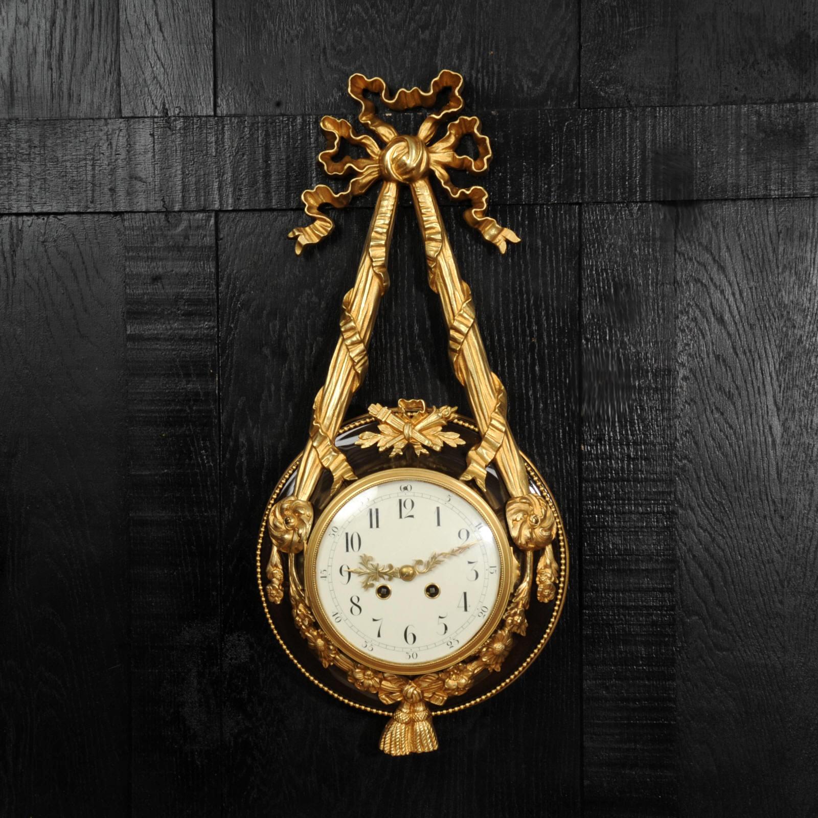 Gran y soberbio reloj de pared de estilo Luis XVI, obra de Samuel Marti. Está formado por un cuerpo redondo de bronce que aloja el movimiento suspendido de un brazalete de cinta de ormolina con una cresta de lazo. El cuerpo de bronce está decorado