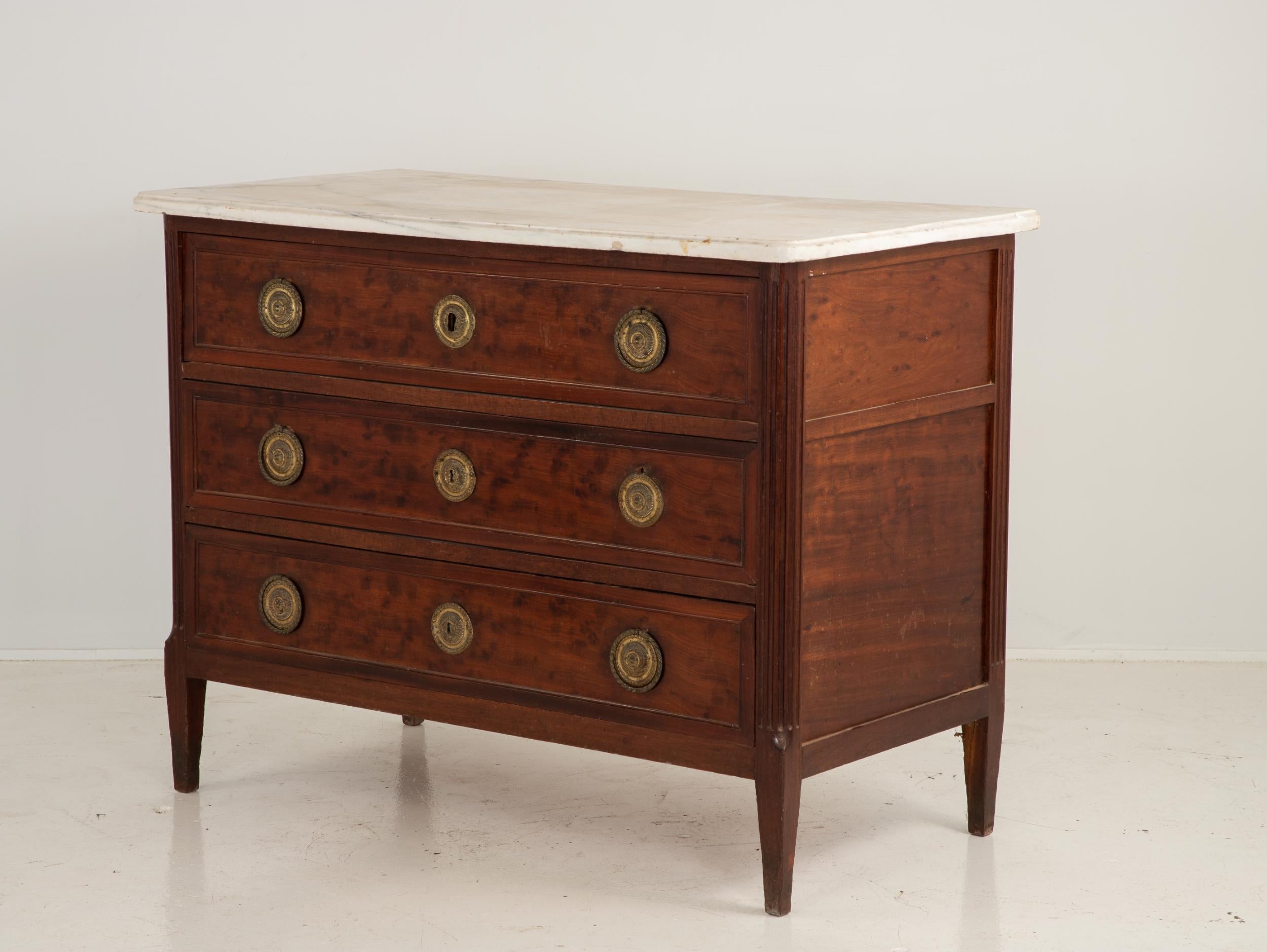 Early 19th century French Louis XVI style chest of drawers with a walnut veneer and topped with white marble. Fluted legs with brass details. Three dovetailed drawers with brass hardware. Minor losses to stone.
    