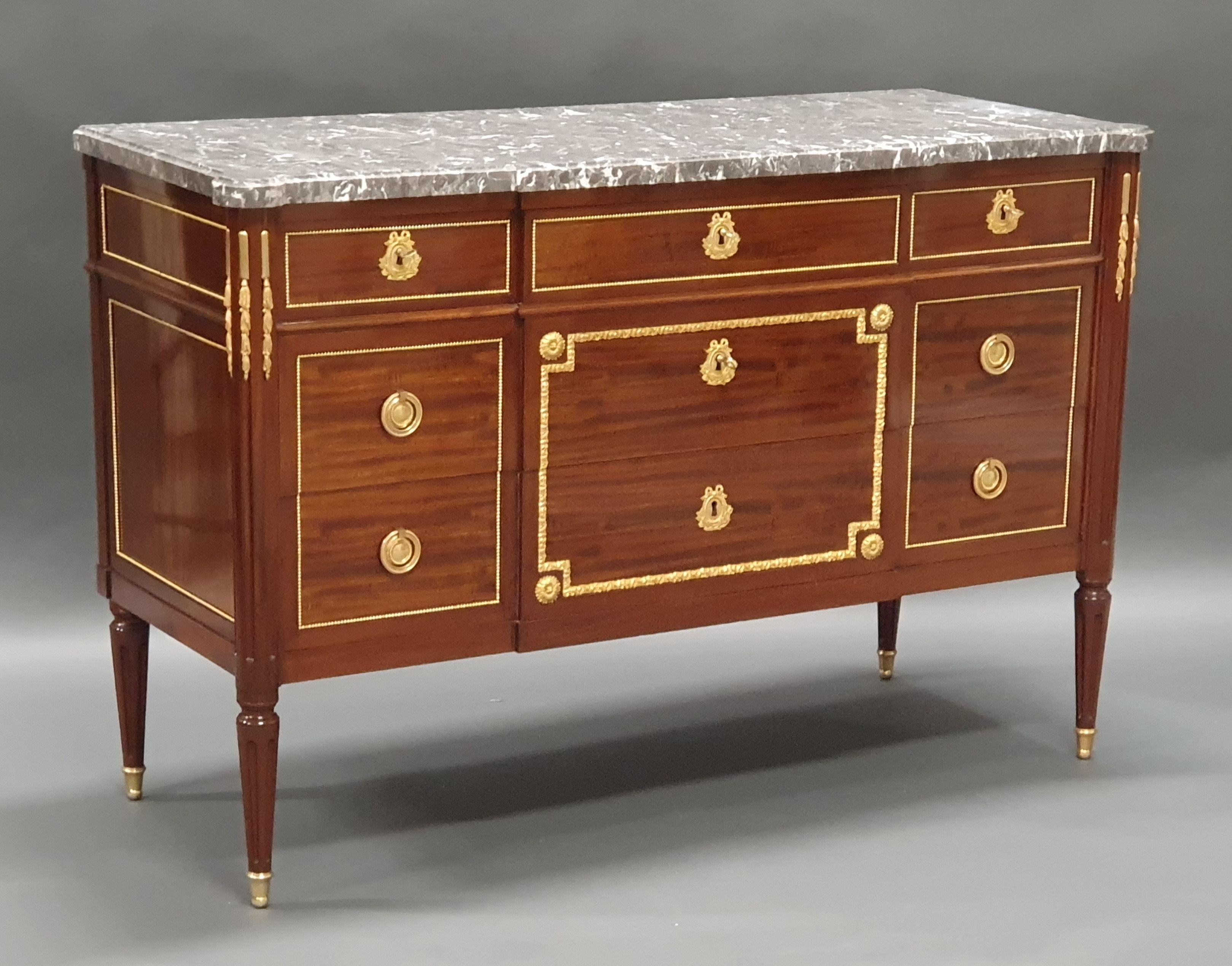 Large Louis XVI Commode in mahogany veneer and rich ornamentation of very finely chiseled gilt bronze, opening five drawers in three rows, central projection and rounded fluted front uprights.

Thick molded Saint Anne gray marble top.

From a