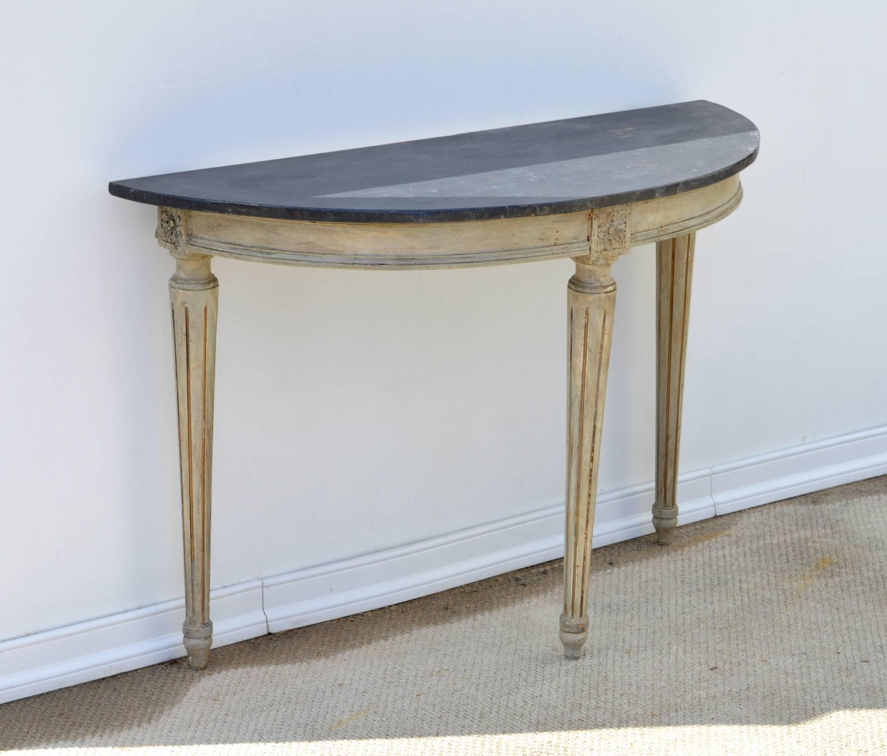 A stunning painted demilune console table in the Louis XVI taste. The worn dark grey wooden top proudly perches atop a simple ogee apron residing on heavily tapered and stop reeded legs. The circa 1960s table has both original and recent layers of