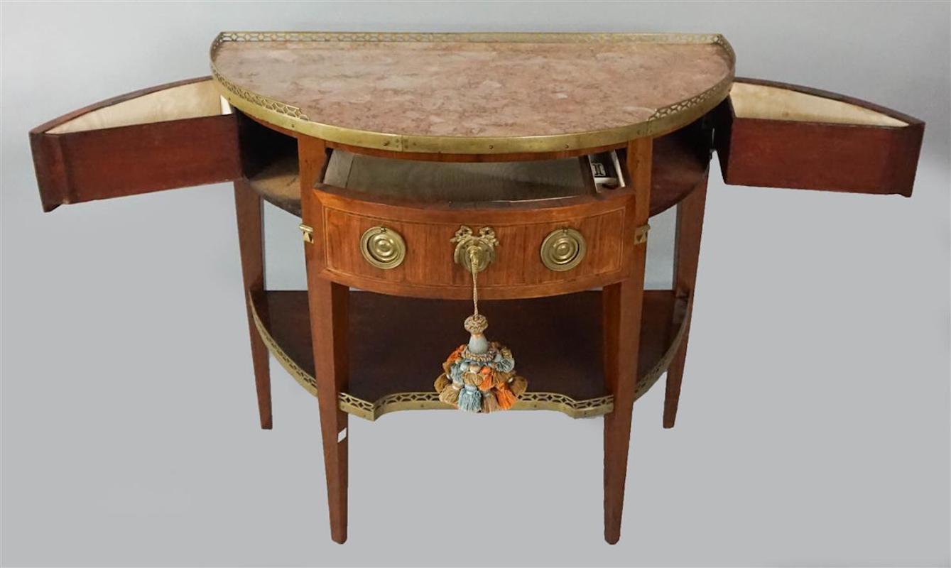 Louis XVI demilune table with pink marble top and gallery. The brass galleried marble demilune top upon a corresponding case with an ormolu decorated center drawer flanked by side drawers over a galleried shelf on squared tapered legs with ormolu