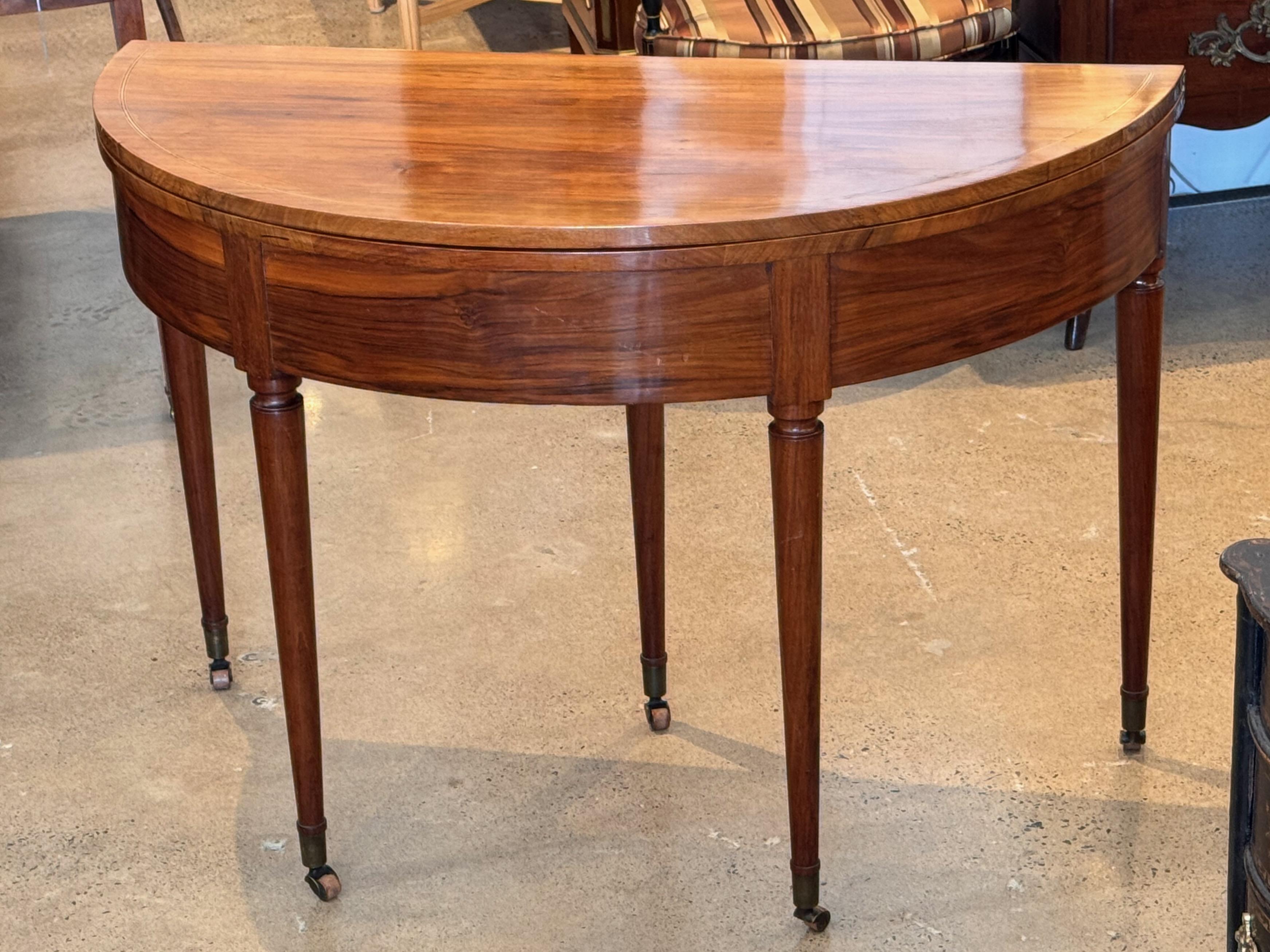 A Louis XVI-style fold-over table with a mahogany top and elegant tapered legs. The table features a demi-lune design that opens to a full circular surface, supported by pull out back leg for stability. A discreet drawer is integrated into the