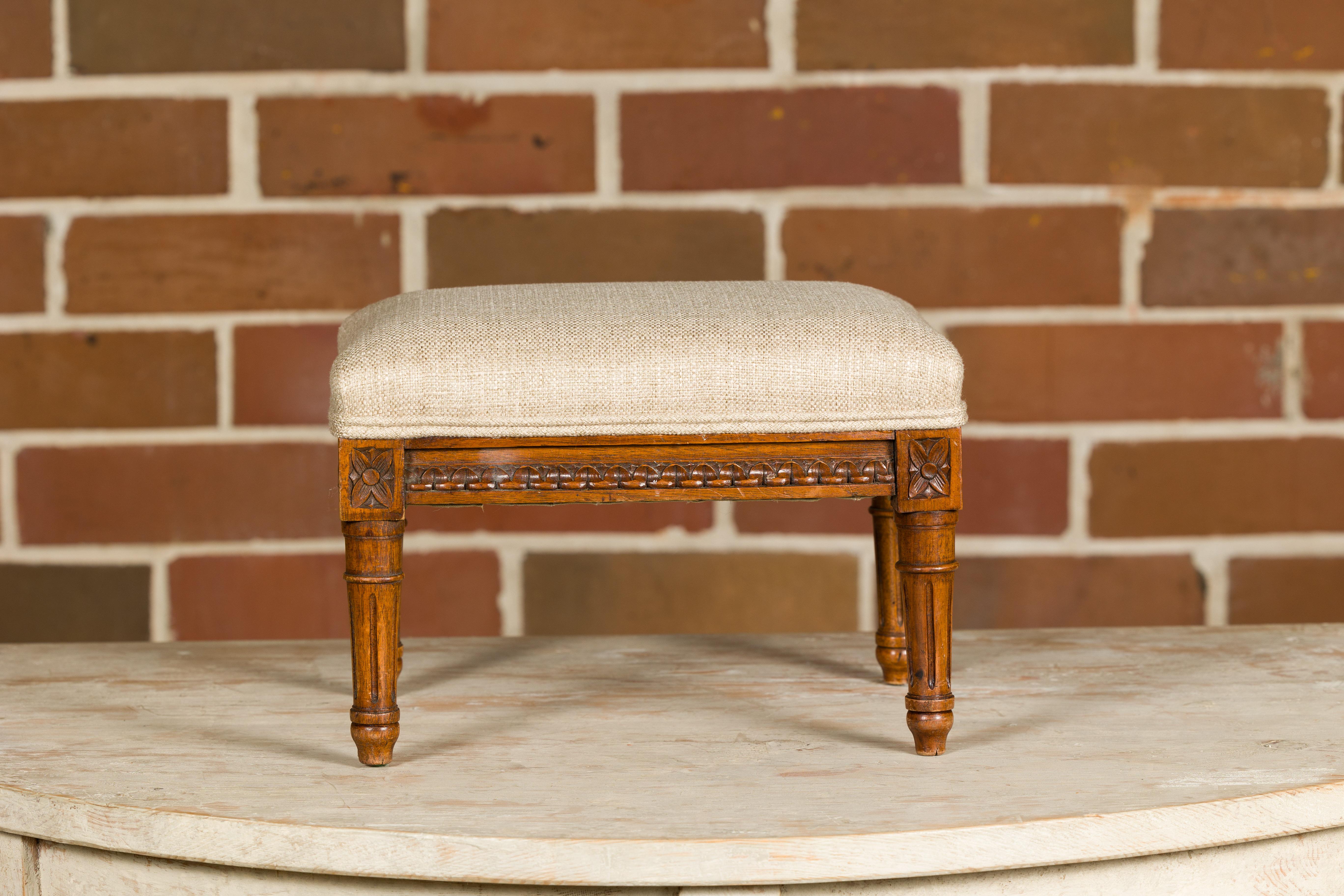 A French Louis XVI style footstool from circa 1900 with carved rais-de-cœur and rosettes motifs on the apron, fluted legs and custom upholstery. This charming French Louis XVI style footstool, dating from around 1900, is a representation of the