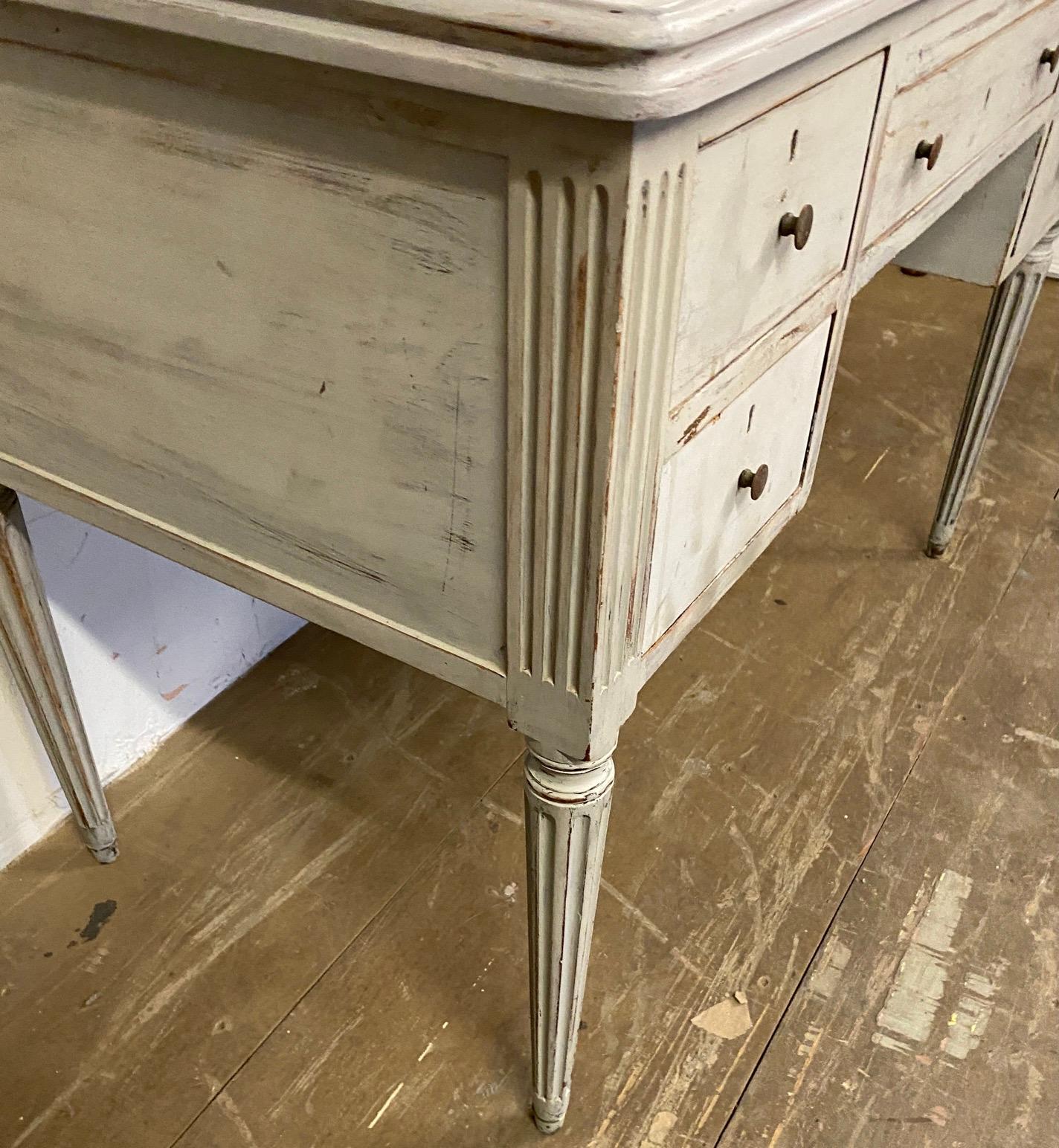This painted rectangular shape Louis XVI style desk stands on four fluted tapered legs. The writing table has two stacked drawers on each side and a drawer in the middle for plenty of storage. Desk is hand painted in a grey palette. The table can