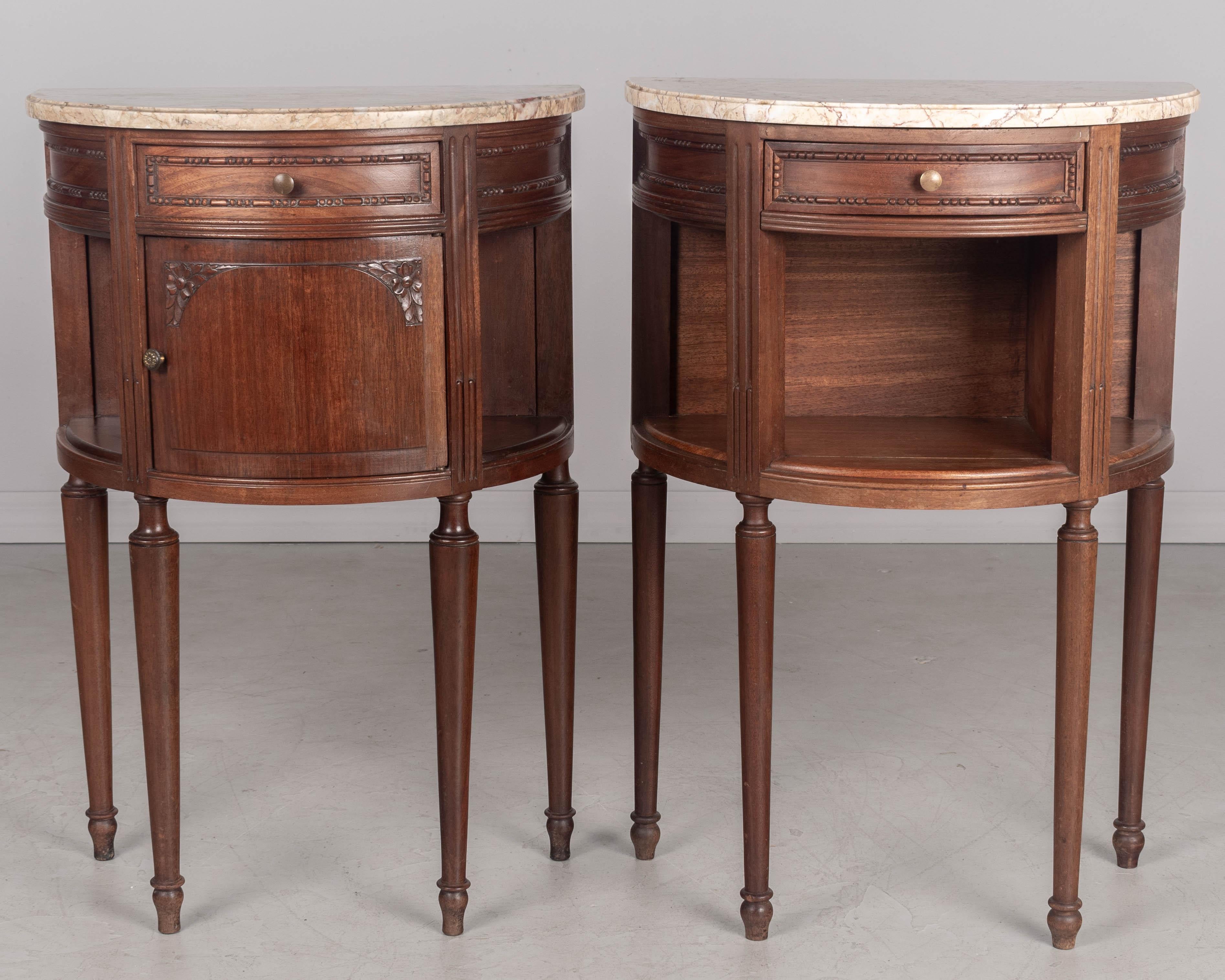 A pair of Louis XVI style French demilune marble top side tables, or nightstands made of solid mahogany, each with a dovetailed drawer and slender turned tapered legs. One stand has a cabinet door with a brass knob and one has an open niche front.