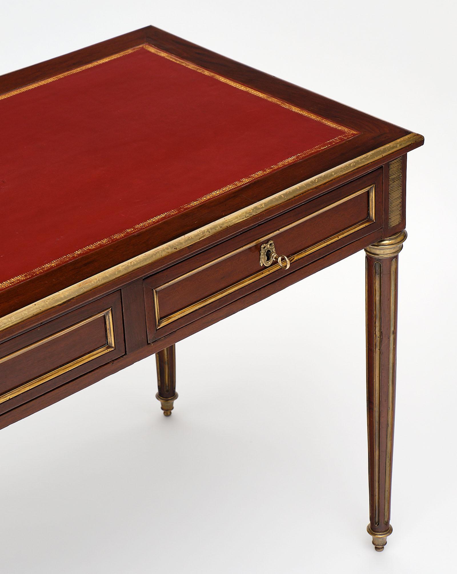French Louis XVI style mahogany desk from the early 20th century, circa 1900. This piece has two drawers with working keys, fluted and tapered legs, brass trim throughout, and its original gold embossed leather top.