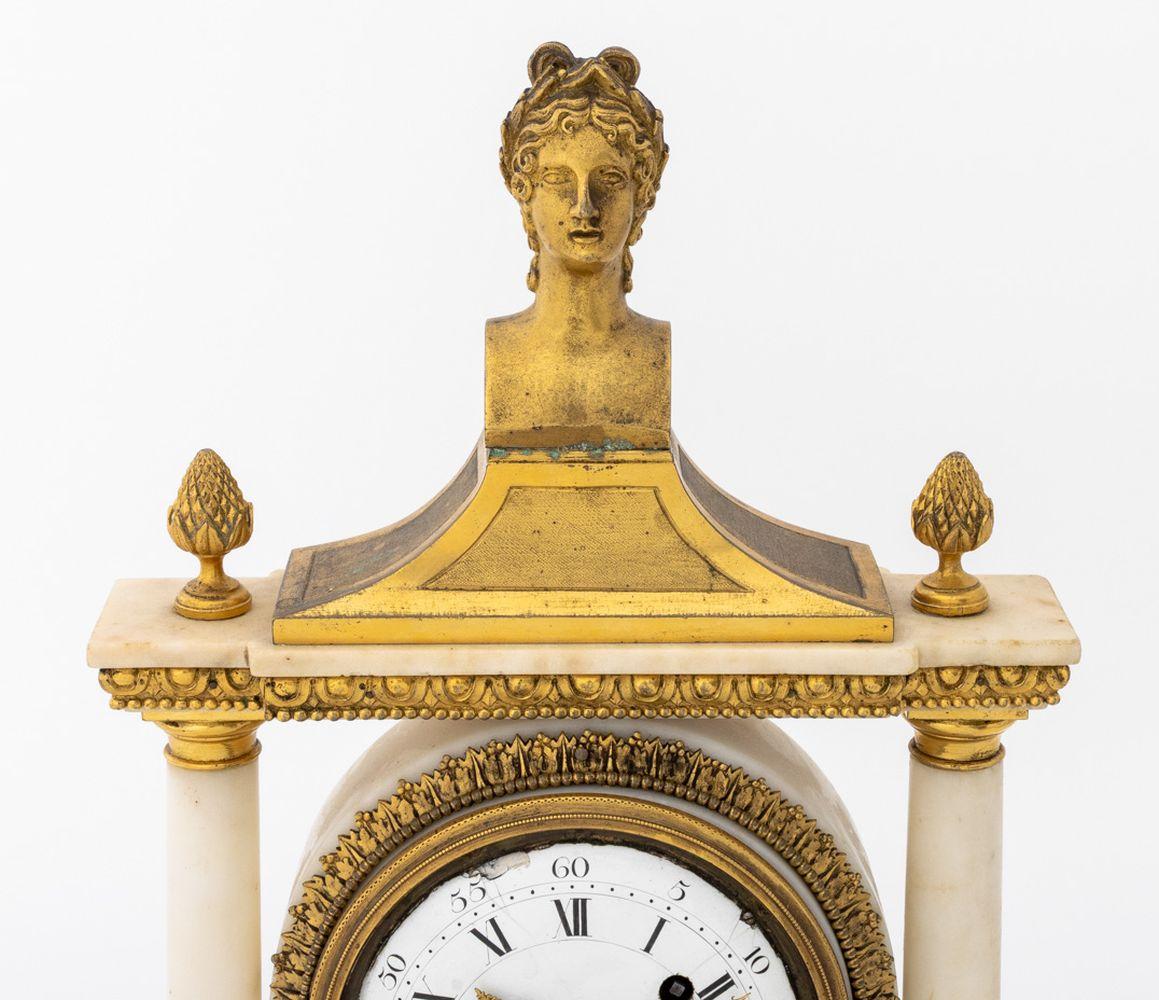 Louis XVI Gilt Bronze and Marble Mantel Clock, late 18th century, of architectural form, the pediment surmounted by an Apollo portrait bust, supporting by tapering columns, centered by the enamel dial, the movement with alarm and strik on bell,