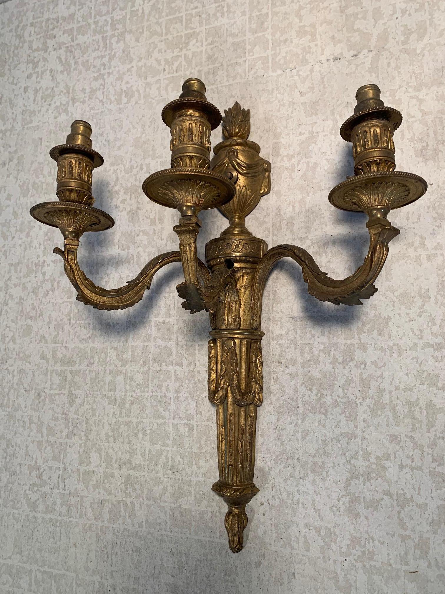 Pair of 3-arm, continental candle sconces with early electrification. Sconces feature foliate sleeved arm with beaded bobesche rim and a tapered fixture body. Since body is surmounted with swaged urn form topped with flame. Original surfaces with