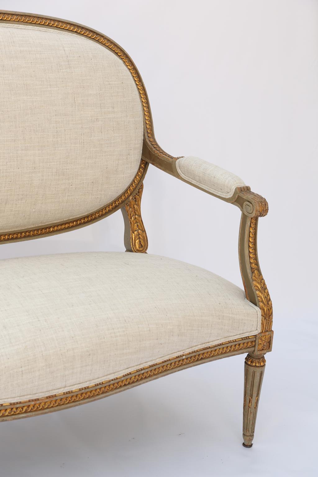 Settee, in Louis XVI style, having a painted and parcel gilt frame, padded medallion backs surmounted by a crest carved as a bow flanked by laureling, joined by acanthine scrolling arms to the padded seat, bowed apron raised on tapering stop-fluted