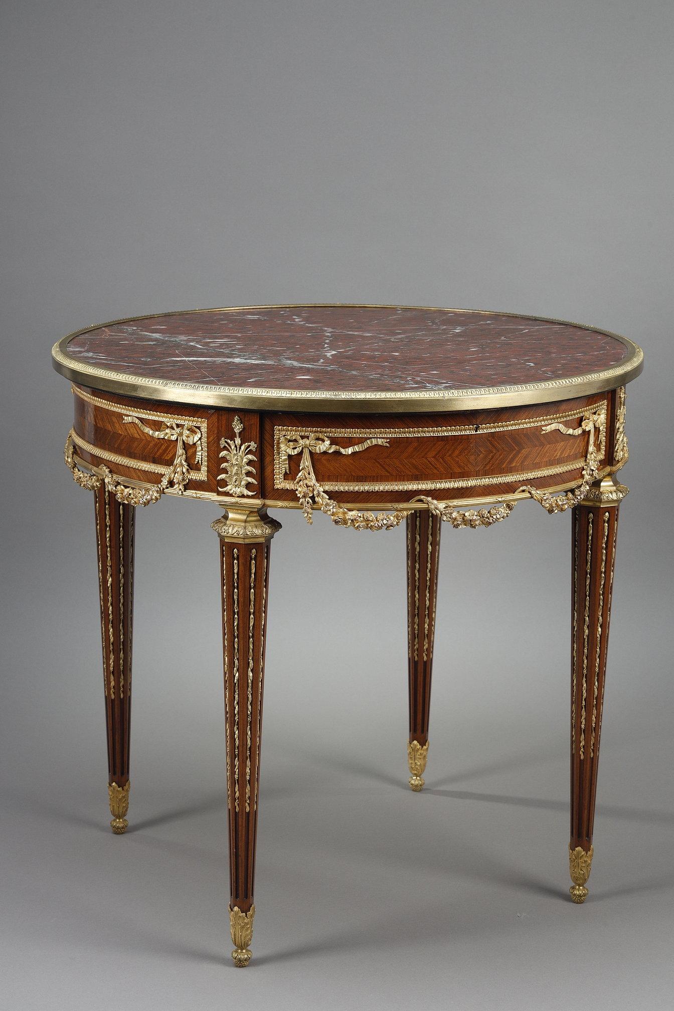 Louis XVI-style pedestal table, remarkable for the elegance of its lines and the quality of its materials. The inlaid precious wood veneer belt is richly adorned with two-tone finely chased gilded bronzes, featuring bows, floral garlands and