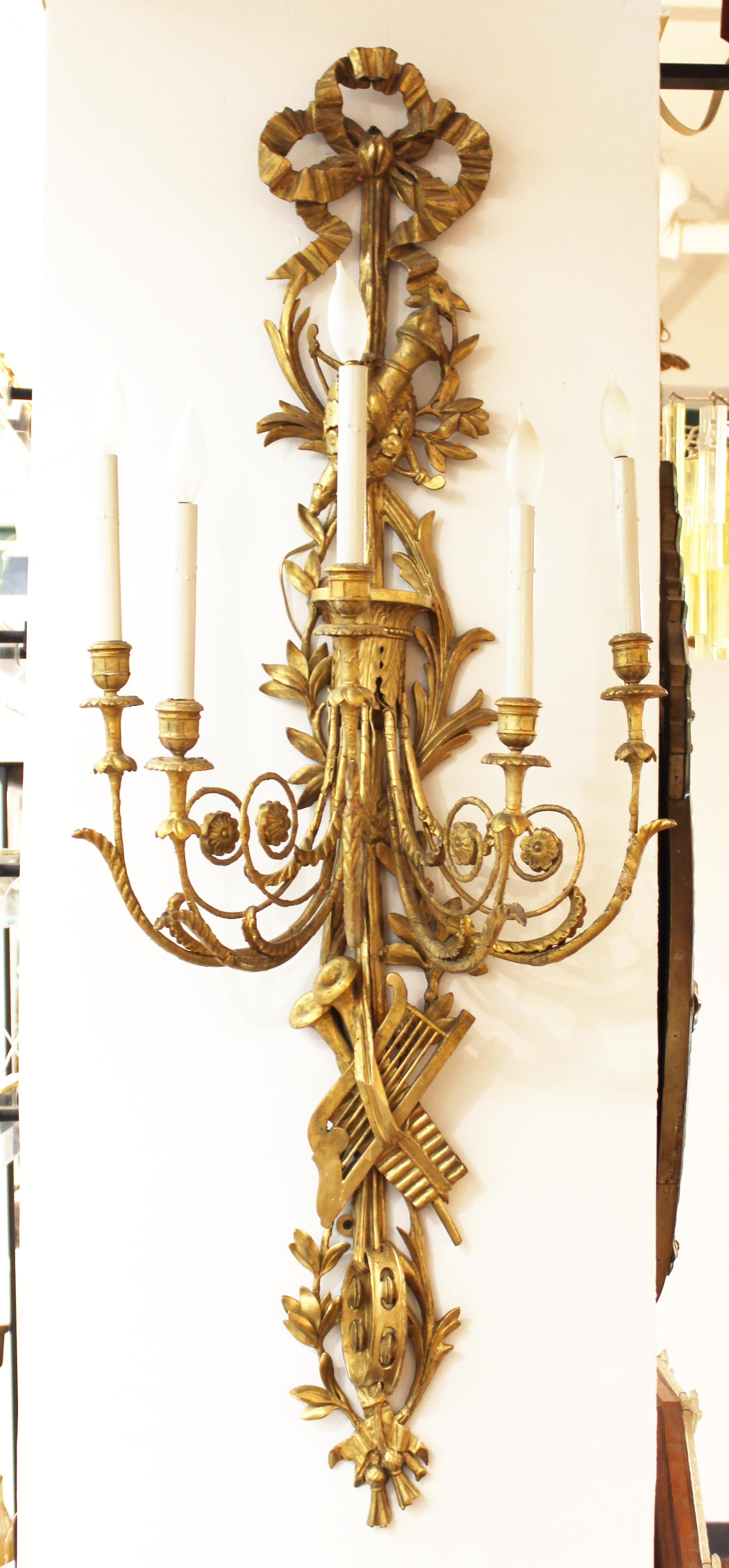 French pair of monumental Louis XVI neoclassical wall sconces. The pair is made of carved and gilt wood and has been electrified. Decorative carved trophies and musical arts elements adorn the upper and lower sections. The pair is mirrored and in