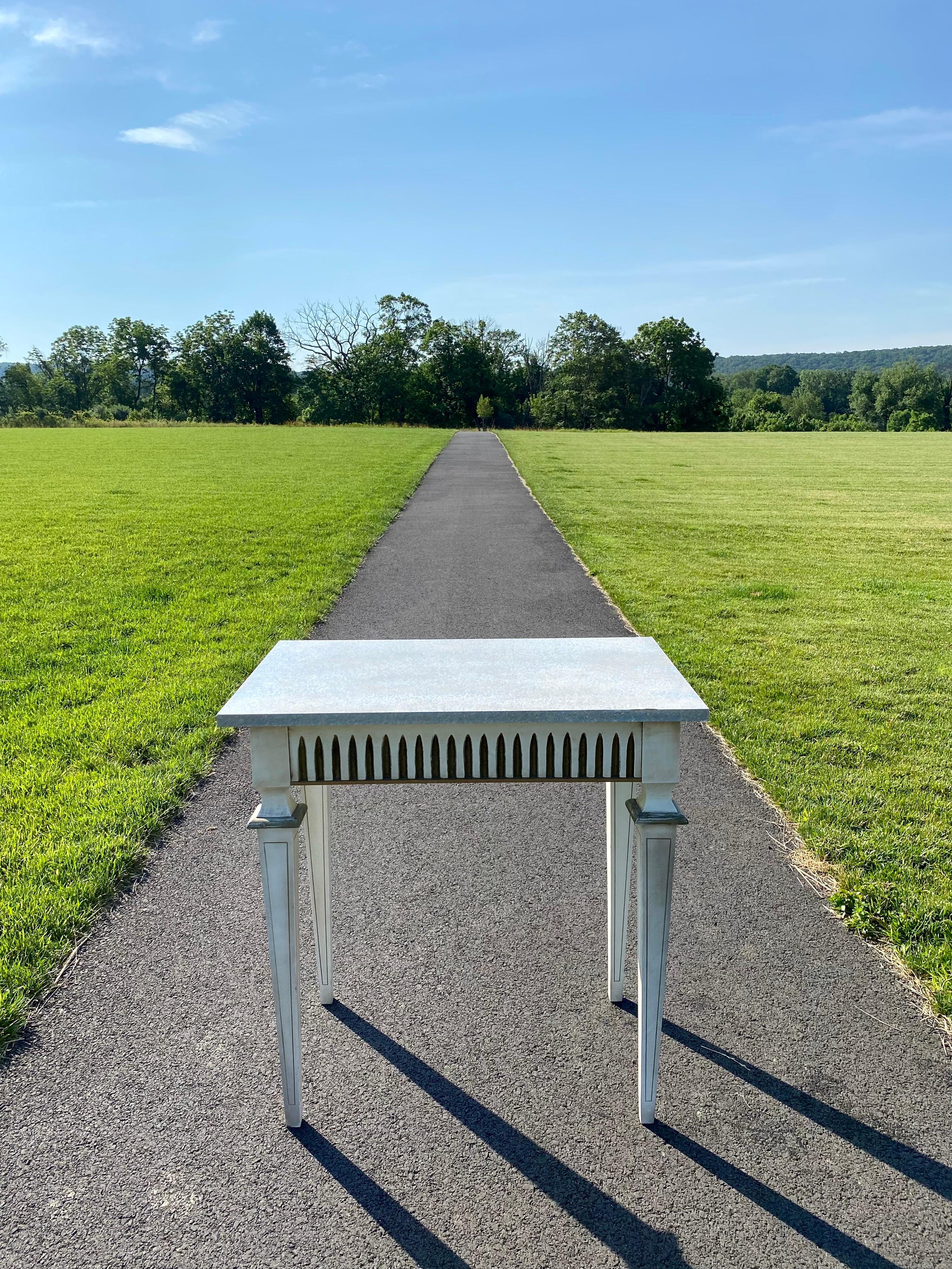 Louis XVI style cream painted wood console table with gilt accents. Apron features fluted carved detailing and tapered legs. Wood top has a textured pale blue-grey painted finish. This Neoclassical style rectangular table is finished on all four