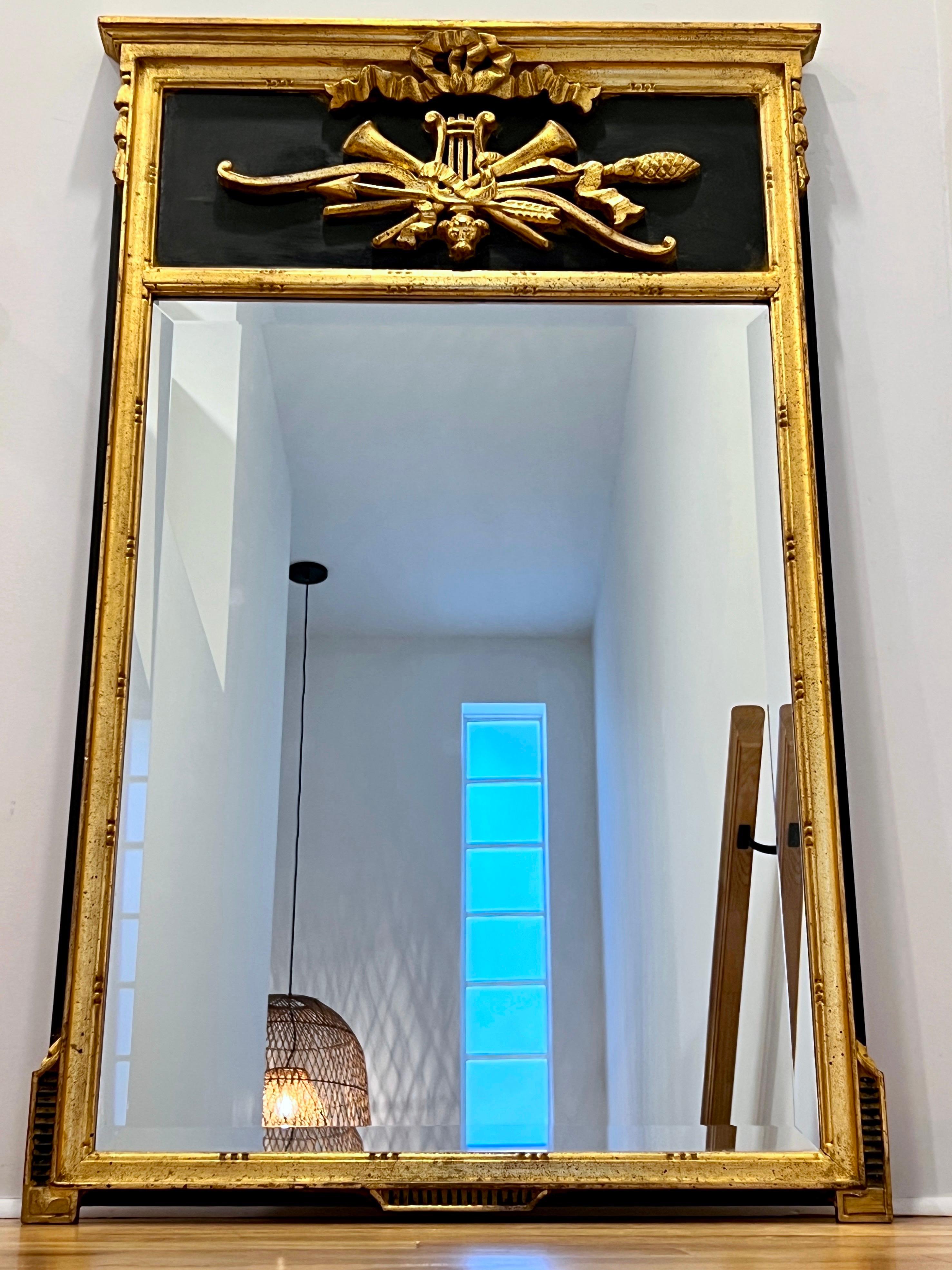 Neoclassical trumeau mirror imported from Italy with hand-carved giltwood frame. The Louis XVI style mirror has a black hand-painted pediment top with ornate motifs in gold leaf gesso. The symbolic motifs feature a lyre, trumpets, bow and arrow,