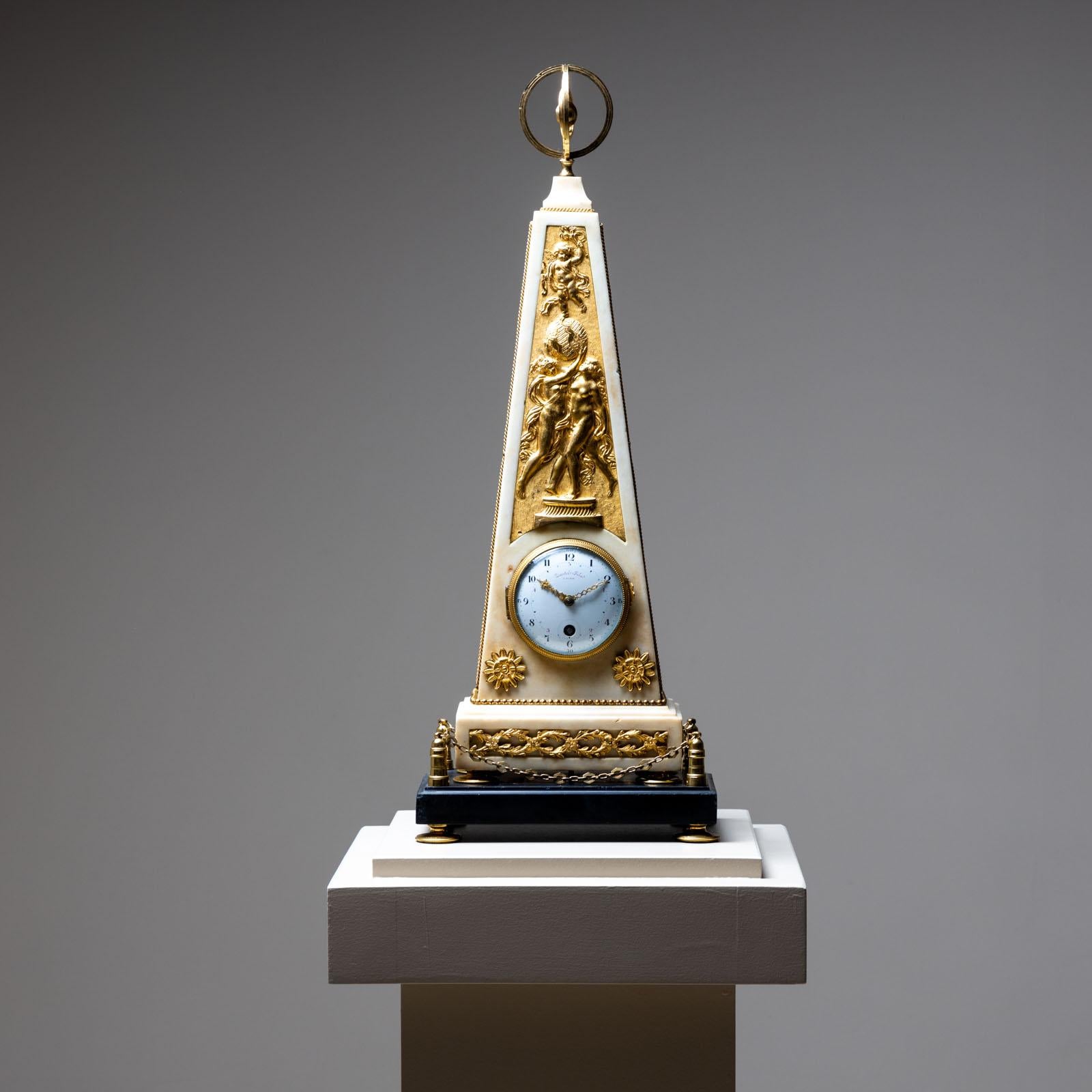 Obelisk-shaped marble clock with fire-gilt bronze appliqués, relief scene, and a surmounting armillary sphere. The clock rests on a black marble plinth with stylized chain railing and low baluster feet. Signed on the dial: “Dautel Fils à Paris.”