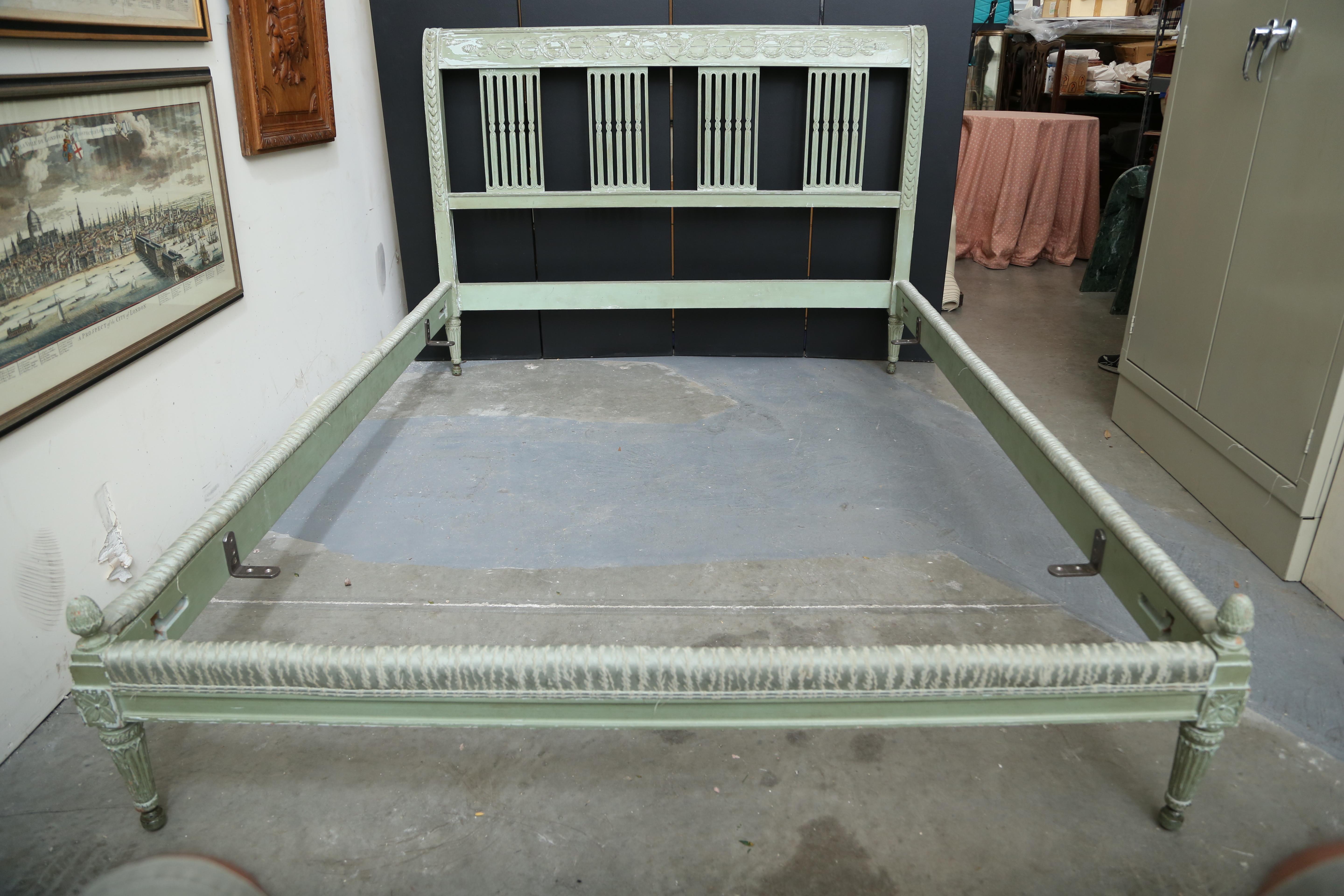 French Louis XVI style painted queen size bed consisting of a headboard, footboard and pair of side rails. The headboards crest rail has a guilloche style carving with laurel leaf swags terminating with pine cones. The foot board has pine cone