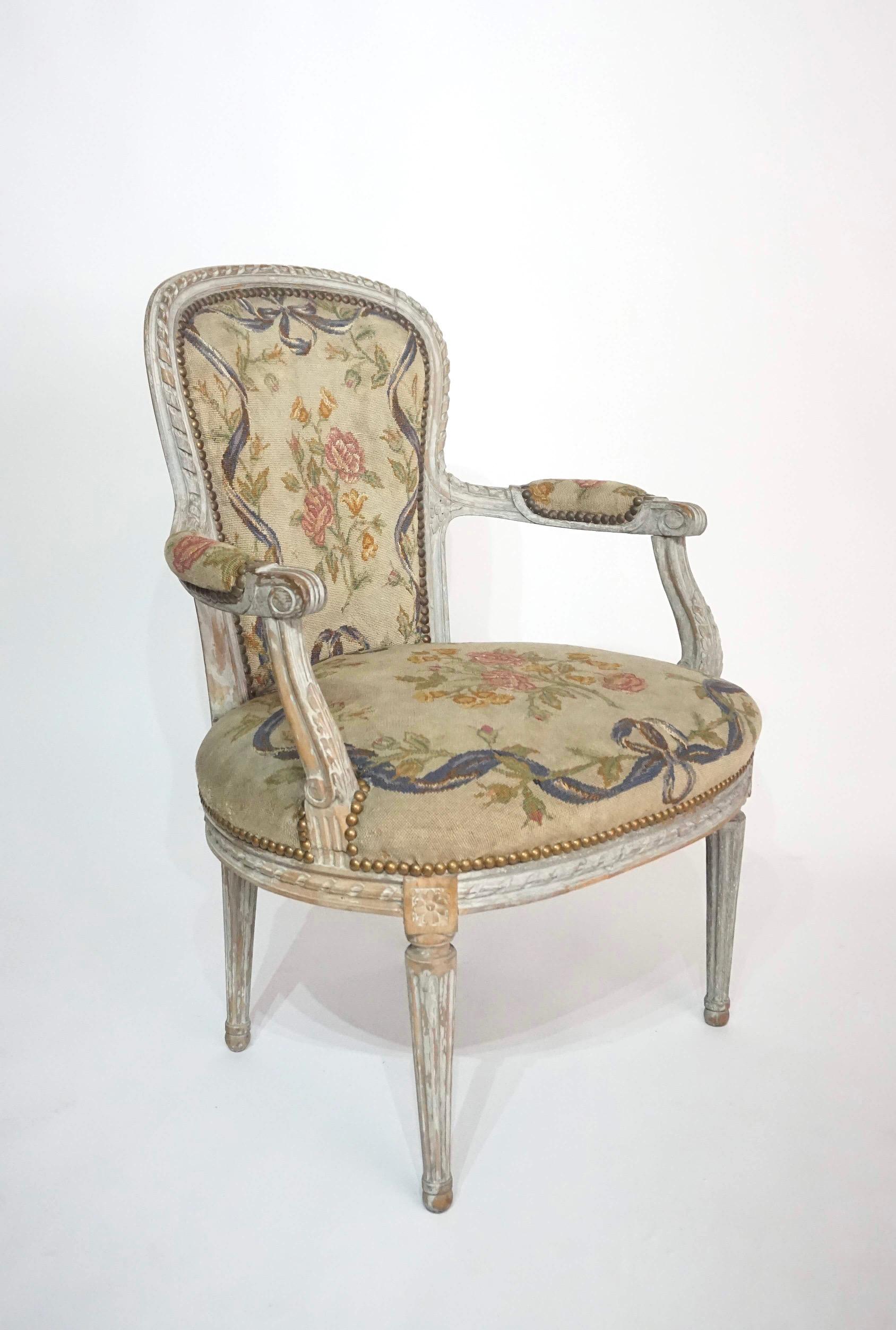 A wonderful circa 1770 French provincial Louis XVI fauteuil in original 'gris' paint, the narrow back and wide seat (typically connoting a ladies chair for gowns) having late 19th century floral and ribbon needlepoint tapestry upholstery.