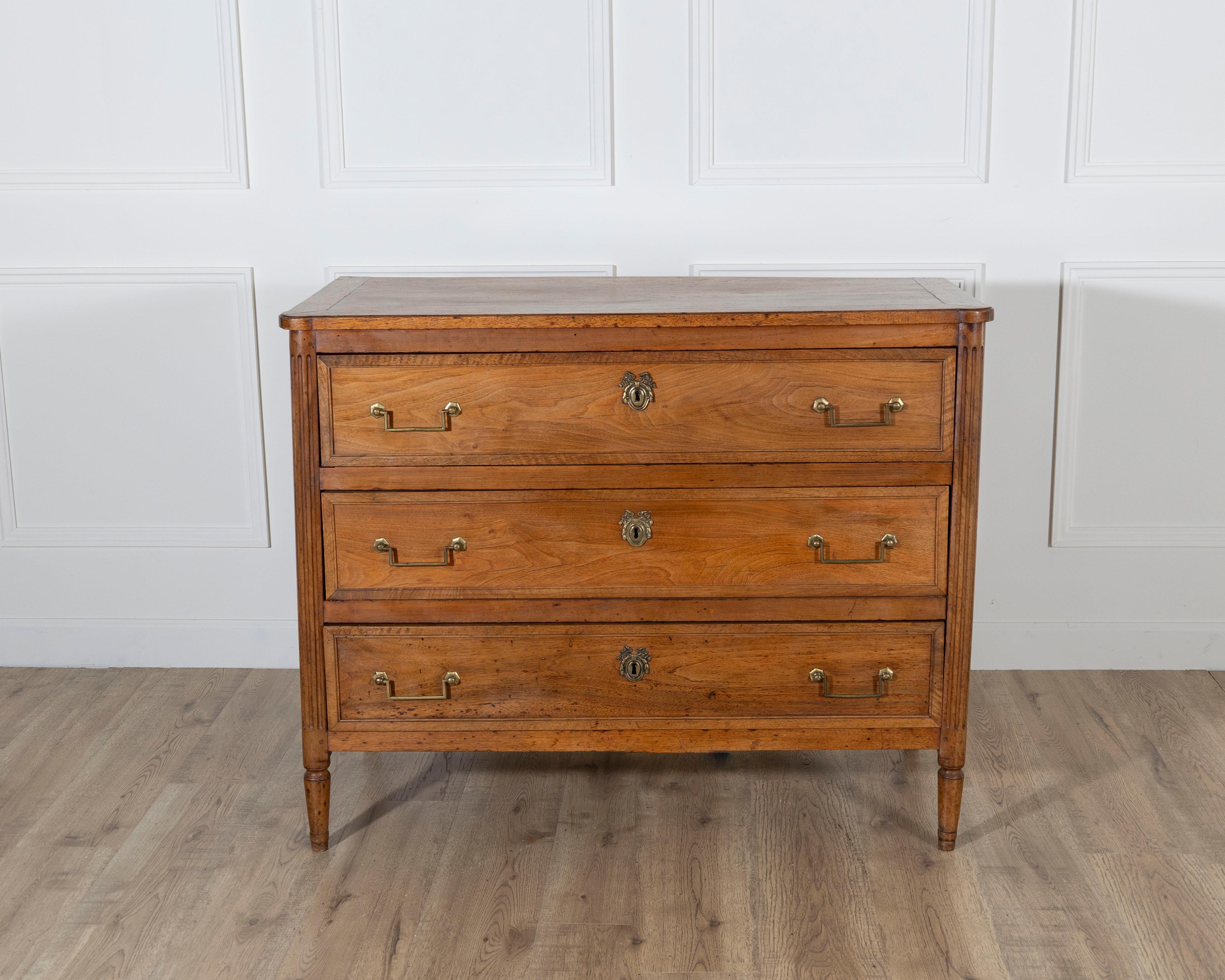 A refined Louis XVI period commode crafted from solid walnut, dating to the late 18th century. This elegant French chest of drawers opens to three full-length drawers, each fitted with original lock escutcheons and classic drop-handle brass pulls,