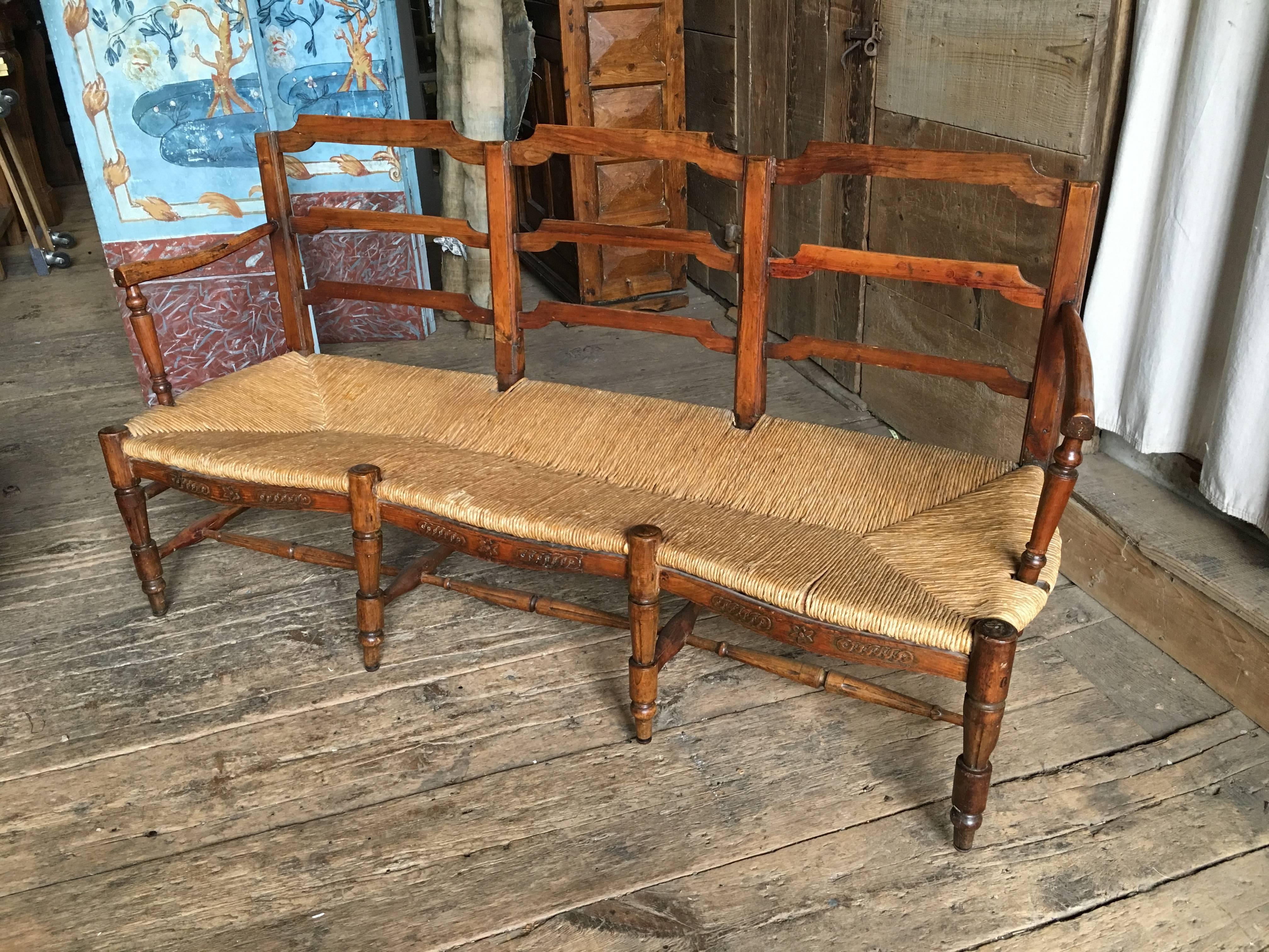 An early 19th century French rush-seat banquette in fruitwood, circa 1800. The back is a simple stylized ladder-back with carved arms supported by turned posts that terminate into the seat. The natural rush seat is supported on eight turned and