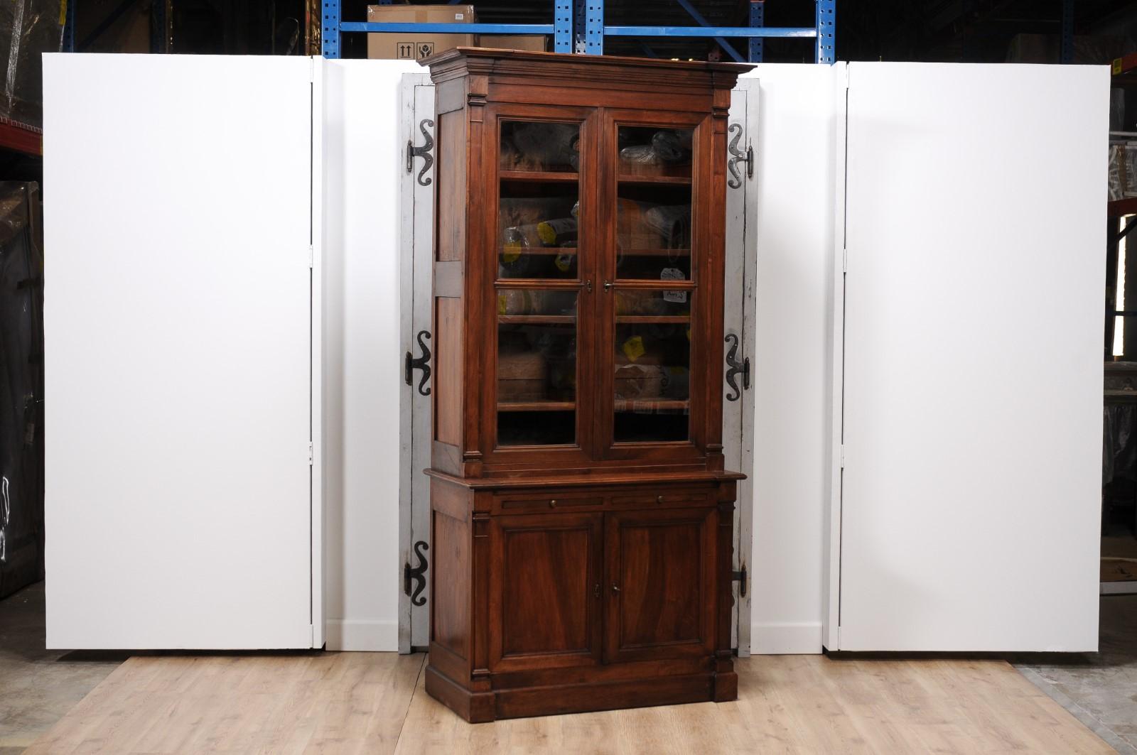 A French Louis XVI style two-part walnut bookcase from circa 1890 with glass doors in the upper section flanked with slender Doric style pilasters, pull-out drawers and wooden doors at the bottom.  Introduce a touch of classic French elegance to