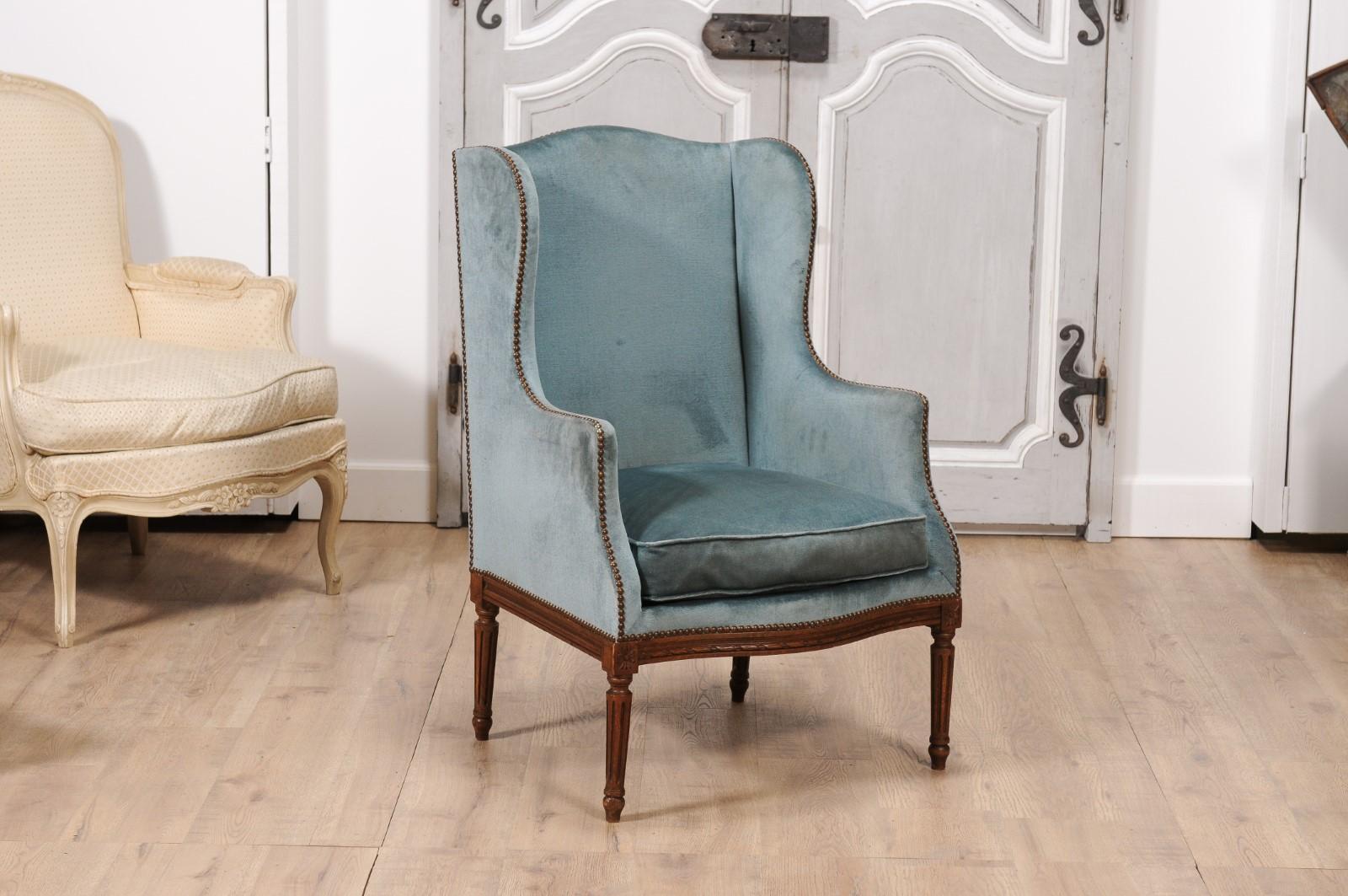 A French Louis XVI style walnut wingback bergère chair from the 19th century with turned fluted legs, carved rosettes on the front knees, twisted rope motif on the apron and blue grey velvet upholstery. Dive into the opulence of 19th-century French