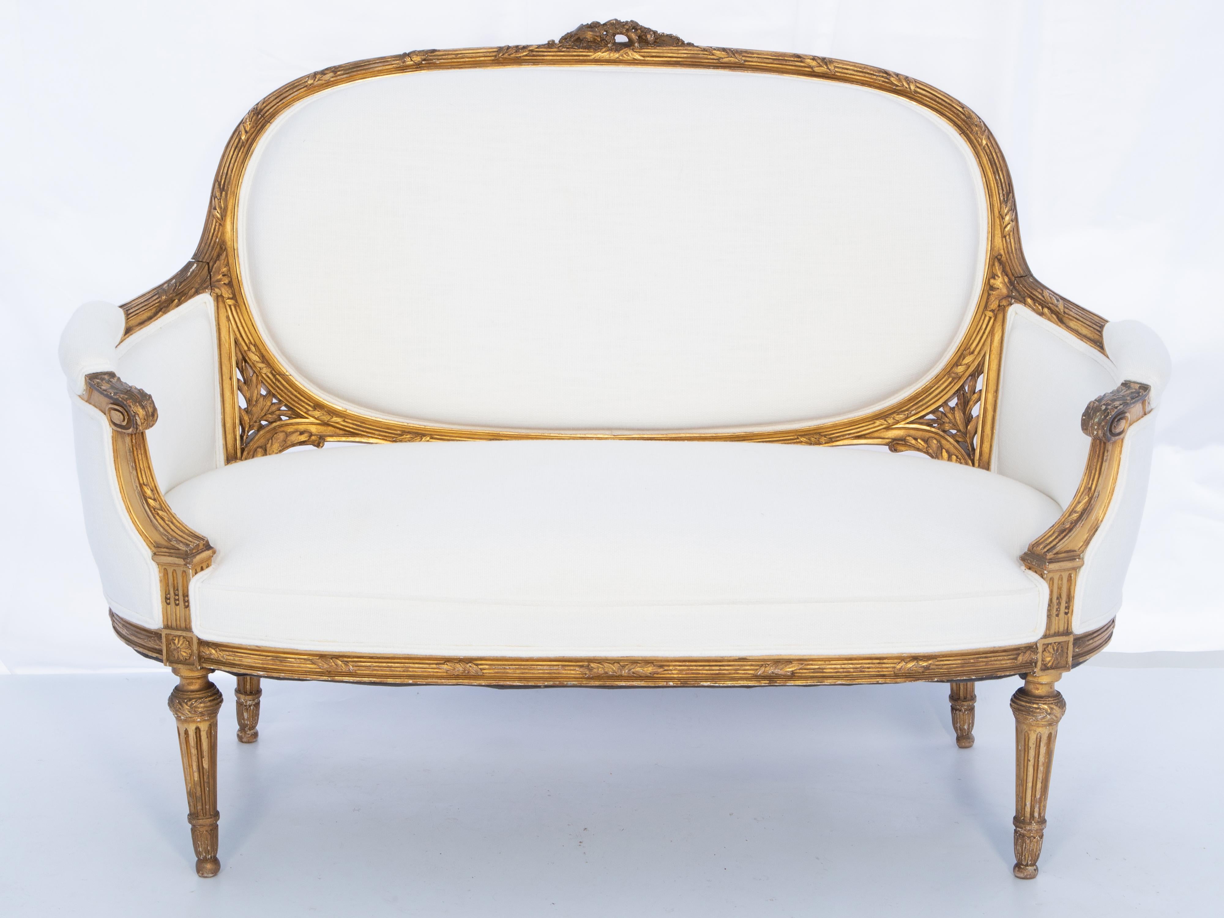 Louis XVI style setee, of carved giltwood, having a reed-and-ribbon carved frame. Its arching crestrail surmounted with a pierced floral ornament, continuing into closed, bergère-style, curved arms with padded elbow rests, ending in foliate-carved