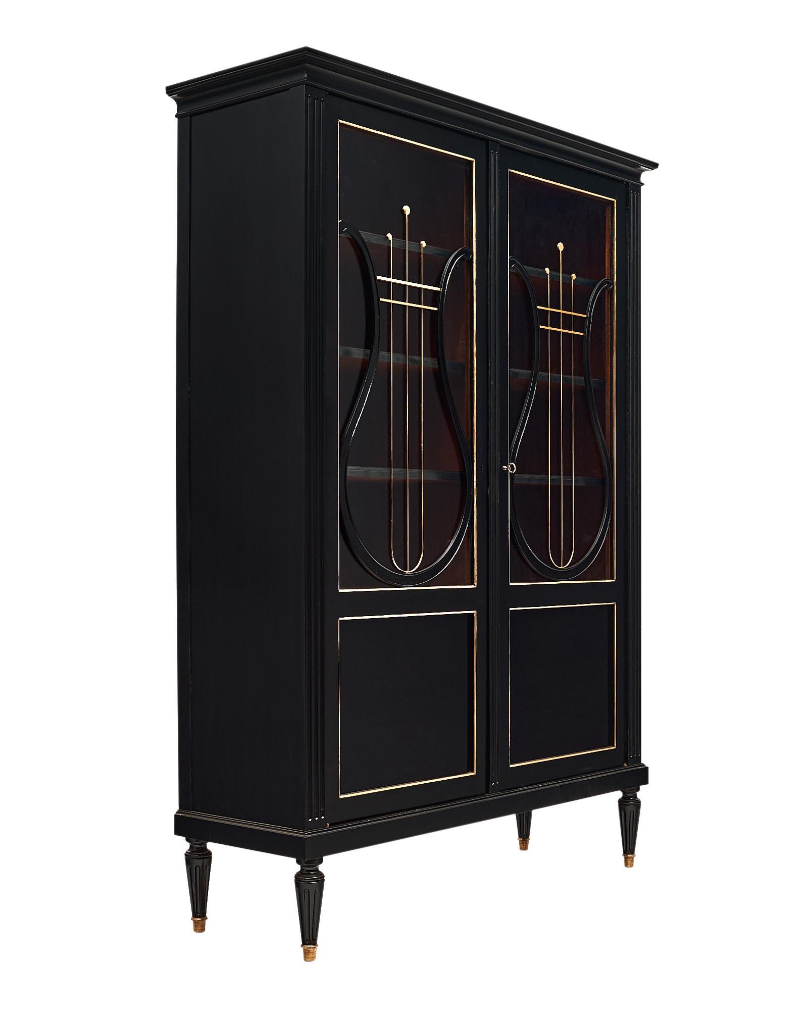 Bookcase “vitrine”, French, made of mahogany that has two glass doors featuring stylized harps. The doors open up to interior shelving. There is gilt brass trim throughout and this piece is finished with an ebonized French polish for luster.
