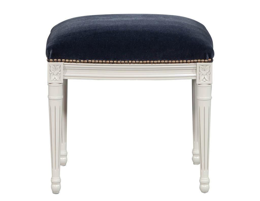 Louis XVI style bench stool, circa 1940s, American. Upholstered in a rich blue velvet with head to head hand applied furniture tack, finished in an antique white glazed finish.