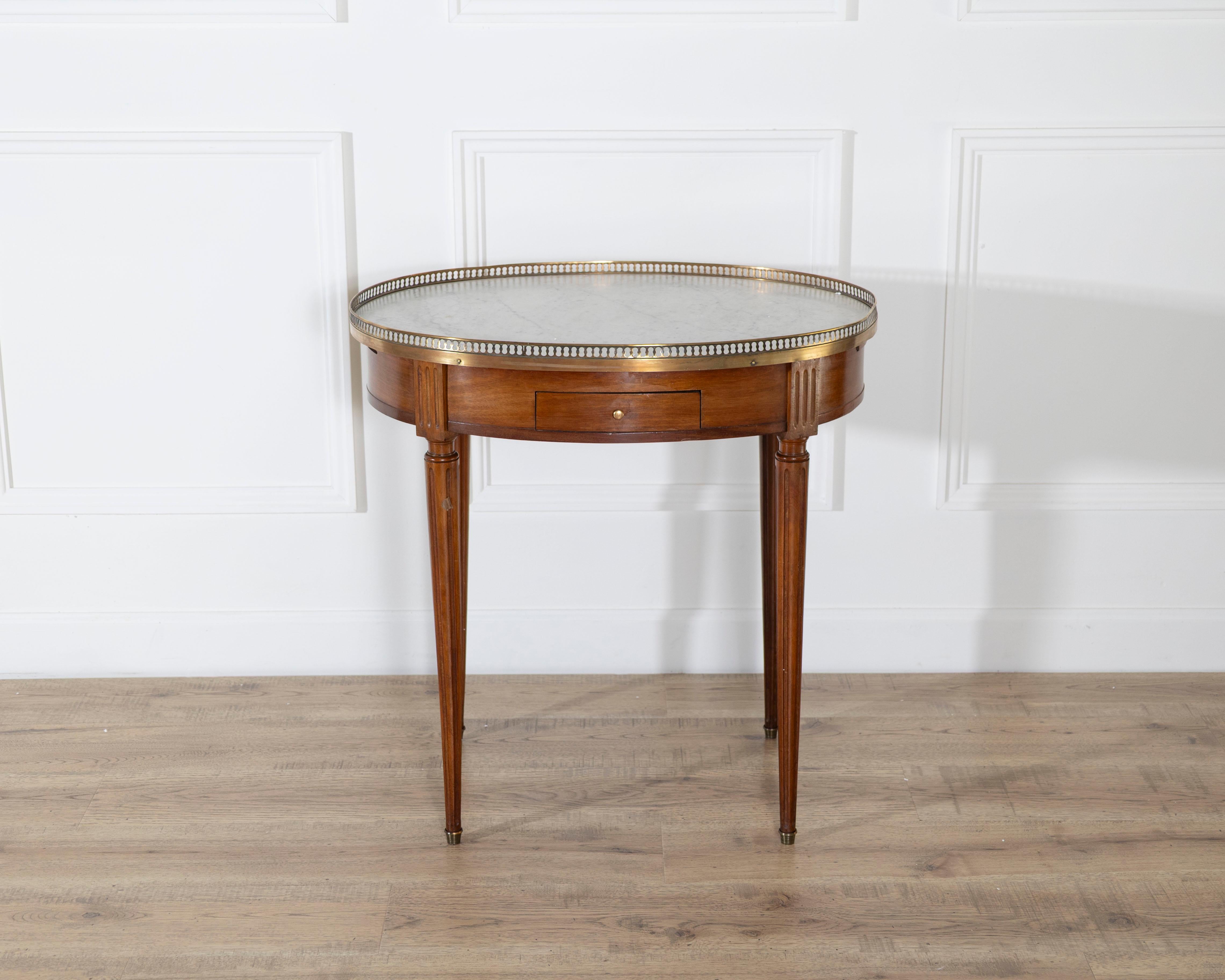 This Louis XVI–style bouillotte table features a circular white marble top bordered by a pierced brass gallery, a defining element of the form. The mahogany frame presents a warm, even tone and includes two small drawers and two sliding candle trays