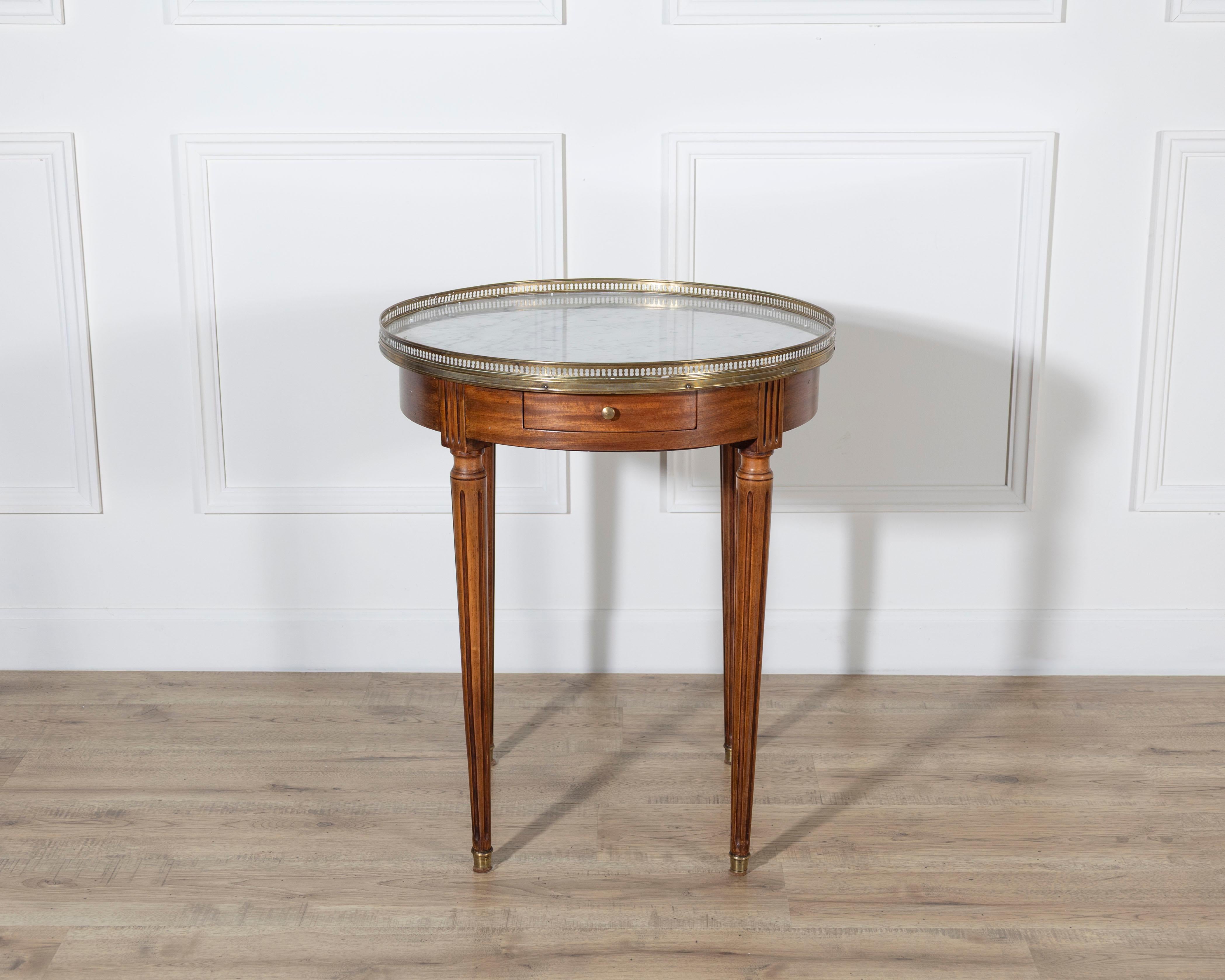 This Louis XVI–style bouillotte table features a circular white marble top bordered by a pierced brass gallery, a defining element of the form. The mahogany frame presents a warm, even tone and includes two small drawers and two sliding candle trays