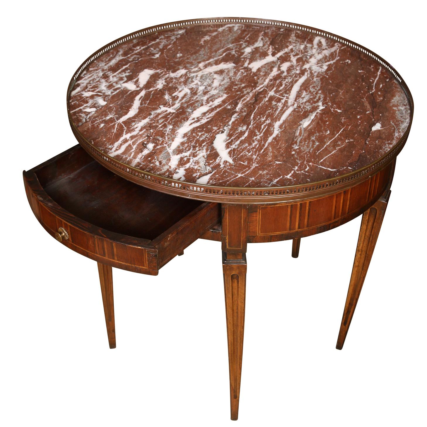 A vintage Louis XVI style bouillotte table with a pierced brass gallery encircling a rouge marble top.  The beautiful gueridon table details include an inlay design on the curved table body and fluted tapered legs with a notch at the top.  An