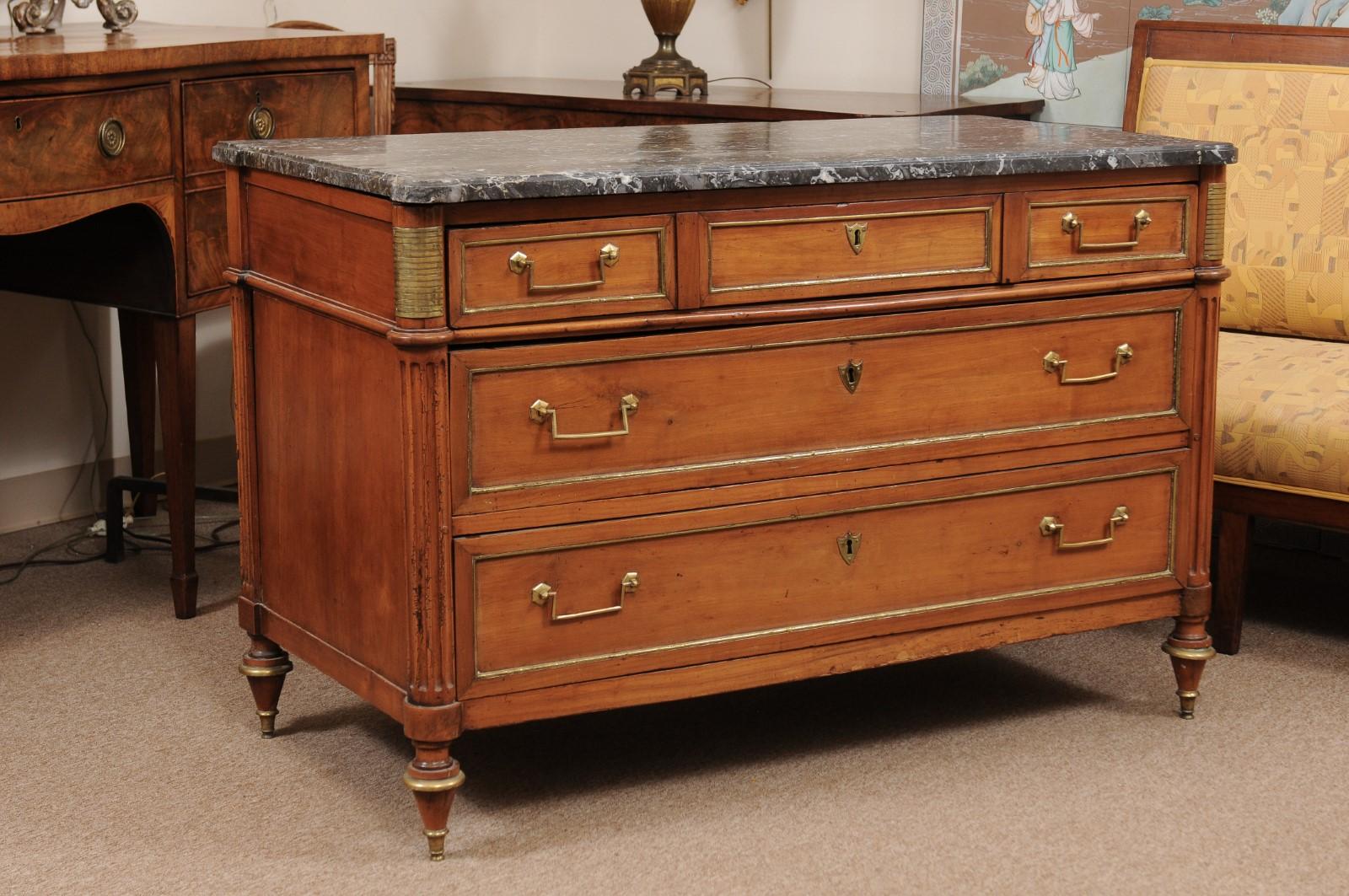A Louis XVI style commode with grey marble top with rounded front corners, 5 drawers below and brass trim. The flanking rounded front corners with carved fluting and brass mounts ending in turned tapered feet.