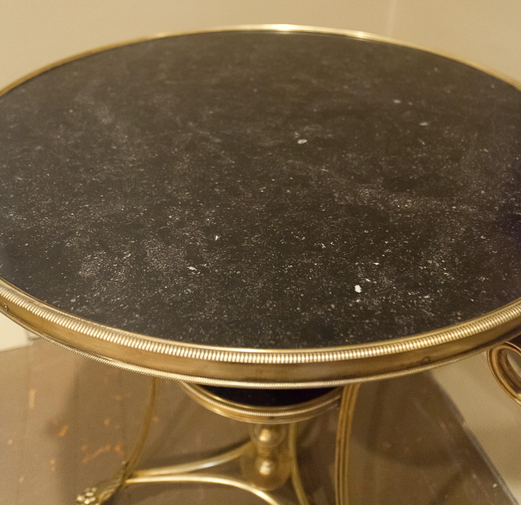 The circular molded fossilized grey marble top of this table is above scroll supports with a hand cast bronze lower tier. On cloven feet with casters joined by a stretcher with an urn in the center. Lower marble shelf is in matching marble. The