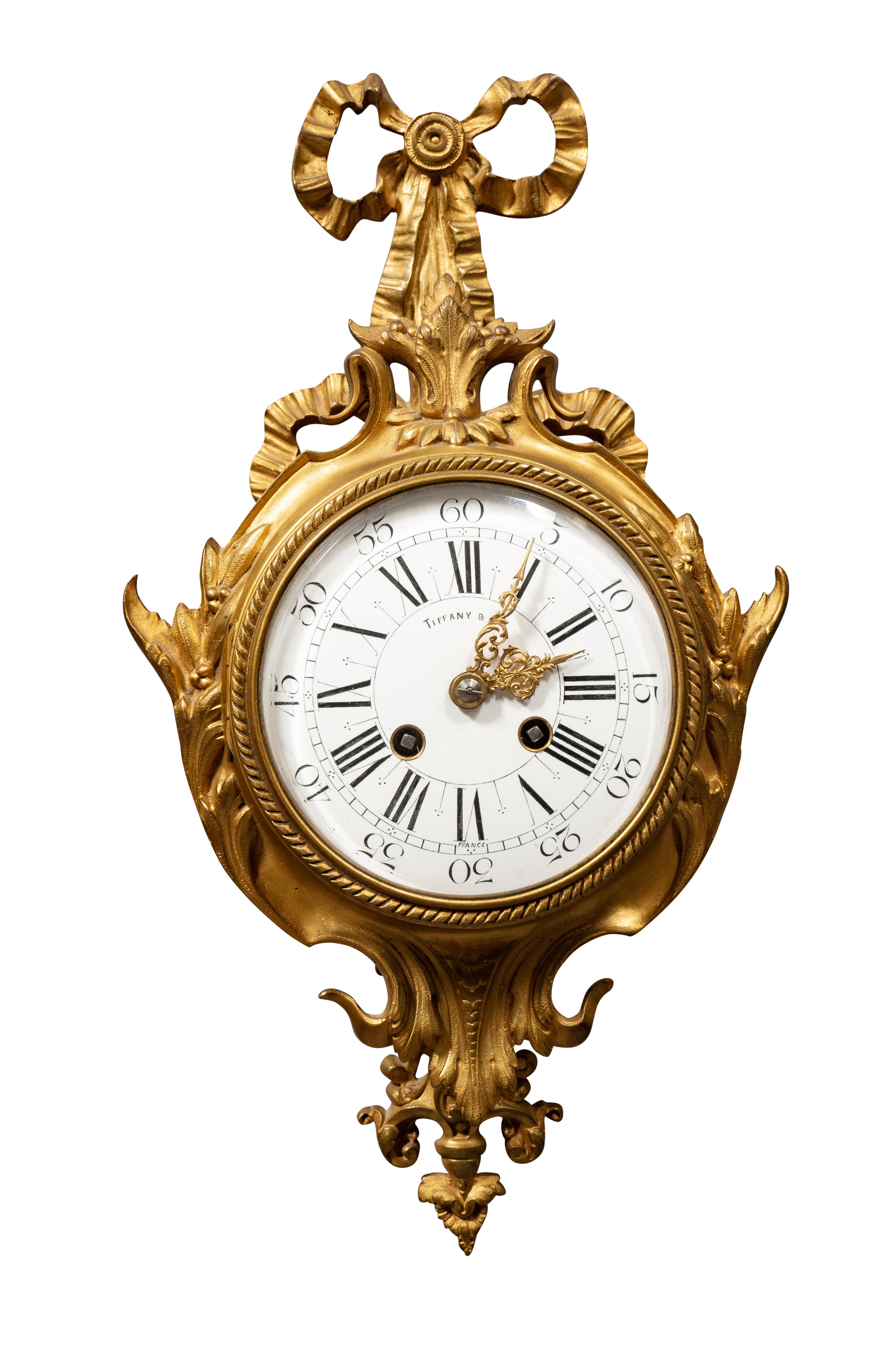 Con un lazo floral sobre una esfera de reloj esmaltada firmada Tiffany & Co. Colocada en una caja de bronce conformada con decoración de hojas y flores.