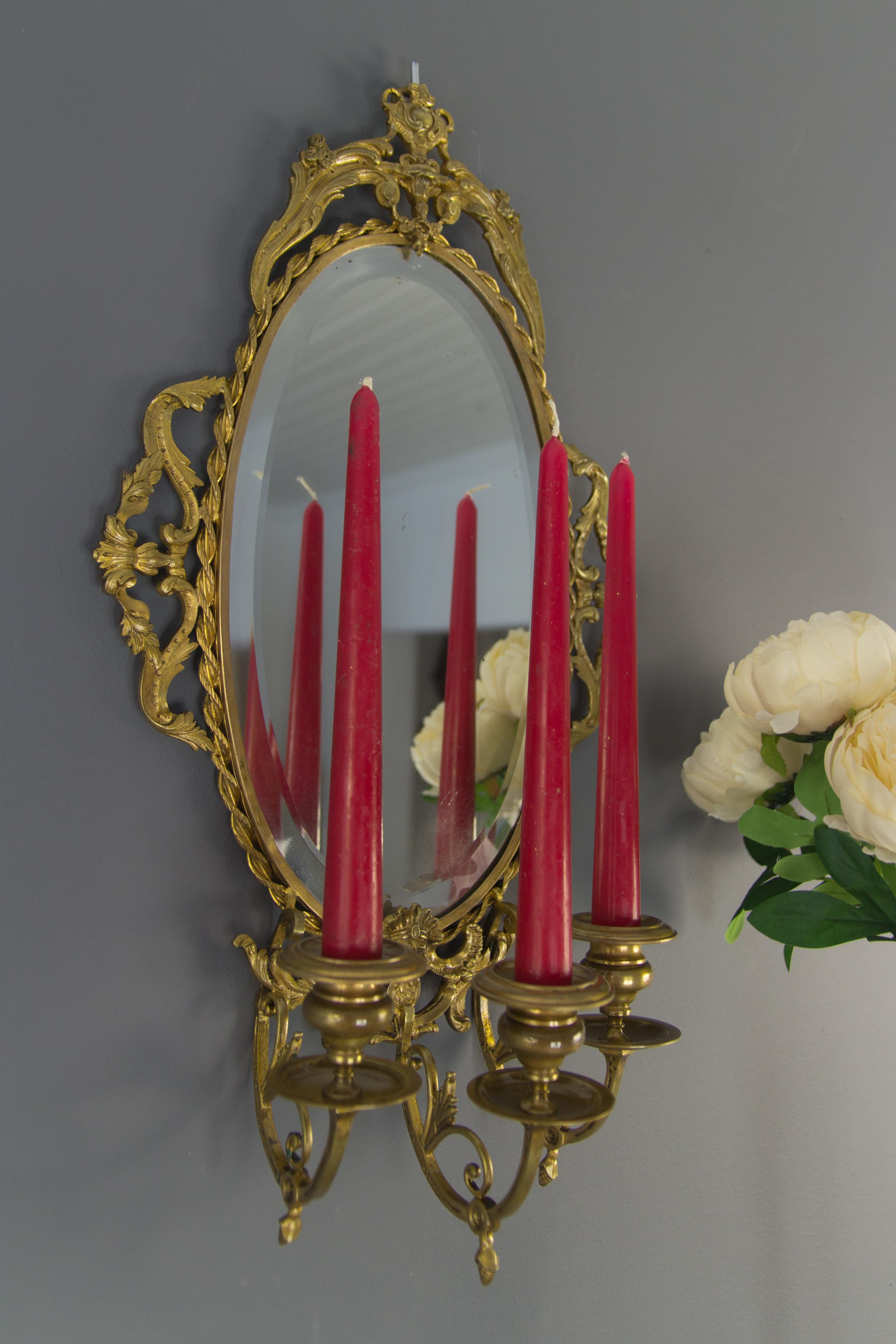 This Louis XVI-style girandole wall mirror has a bronze frame surrounding a beveled oval mirror plate. The frame is decorated with leaves, flowers, scrolls, and a boy’s face. Beneath are three candle sconces, and the arms are decorated with leaf