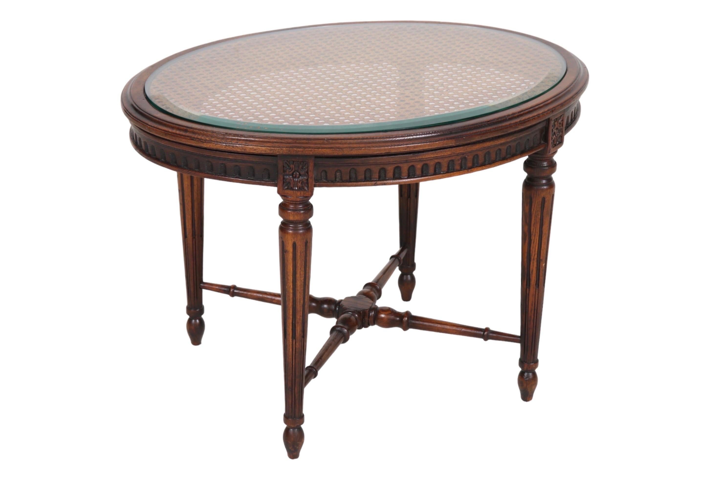 A Louis XVI style oval accent table. The caned tabletop is covered with a beveled glass top. Round tapered legs are fluted, topped with rosettes carved in squares, and finish in turnip feet. Legs are supported with a turned x-stretcher.
