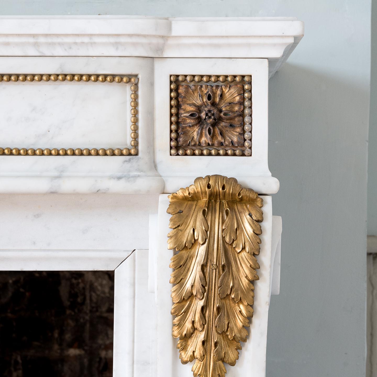 A Louis XVI style Carrara marble and ormolu mounted fireplace, the shaped and moulded shelf above frieze centred by fluted panel adorned with trails of foliage and ribbons, the paterae endblocks with beaded border raised on acanthus clasped scrolled